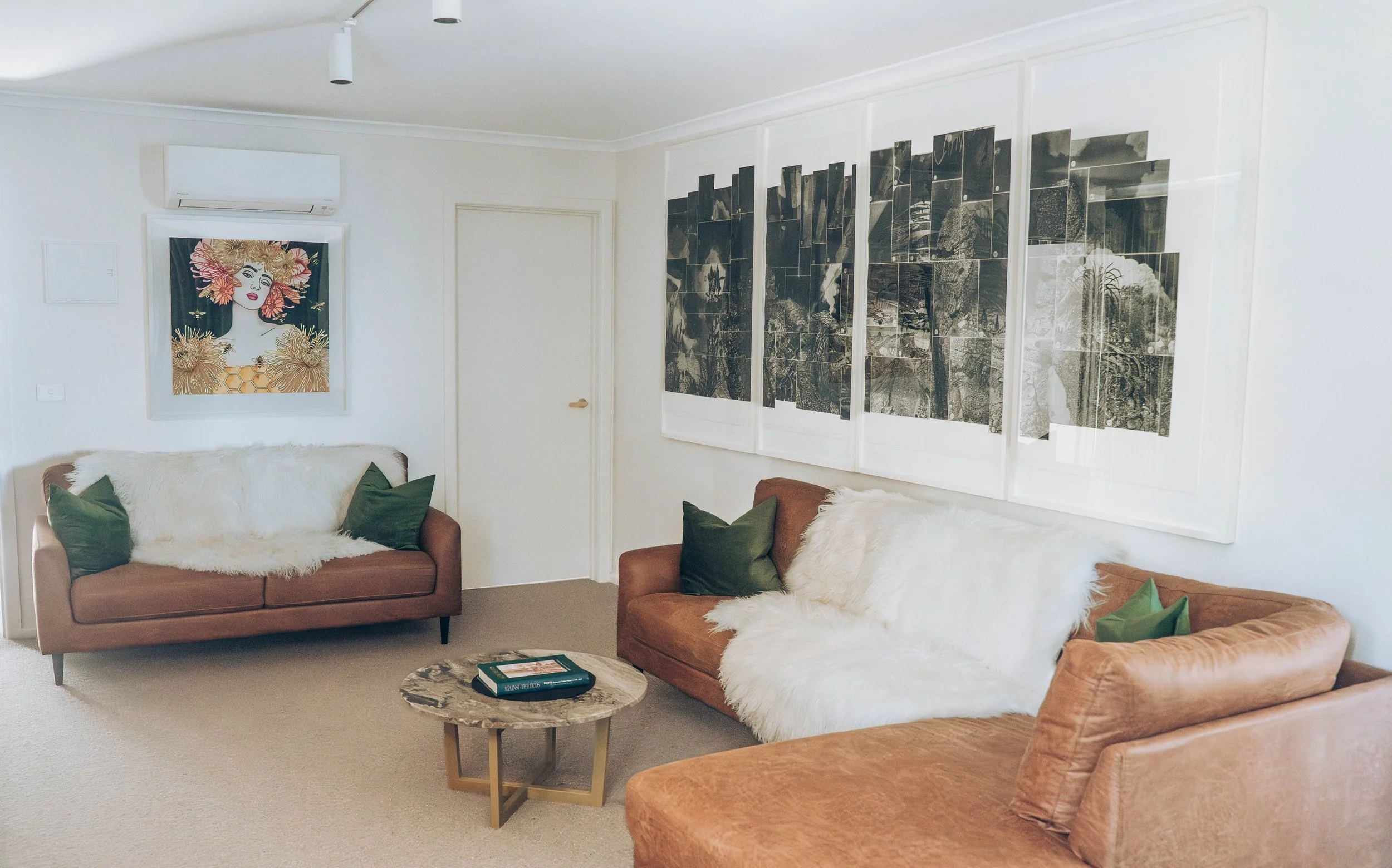 Living room with three sofas, decorative pillows, coffee table with books, wall art, and an air conditioning unit.