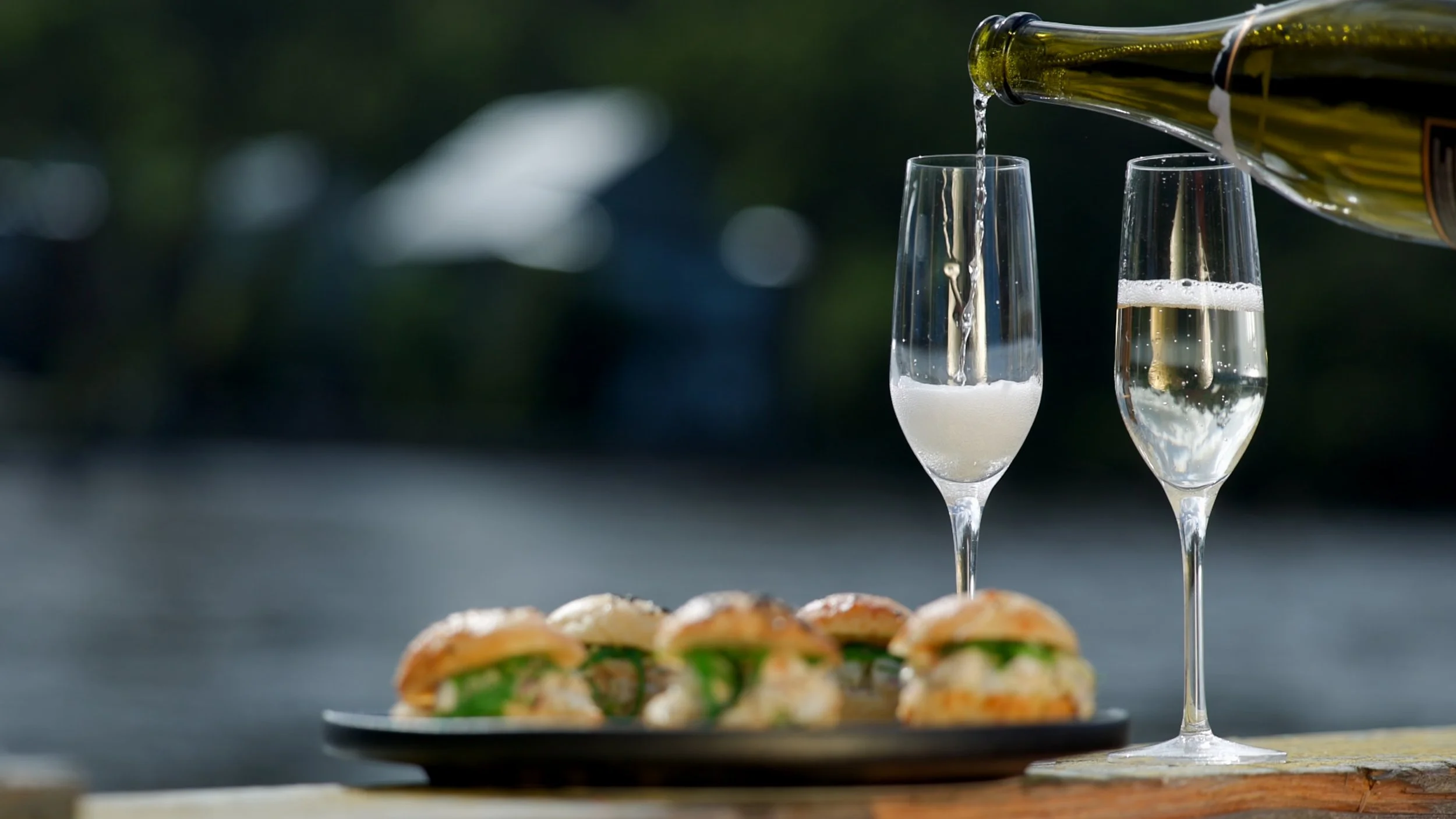 Pouring sparkling wine into two tall champagne flutes with a platter of crayfish sliders in the foreground, outdoor setting with blurred greenery in the background.