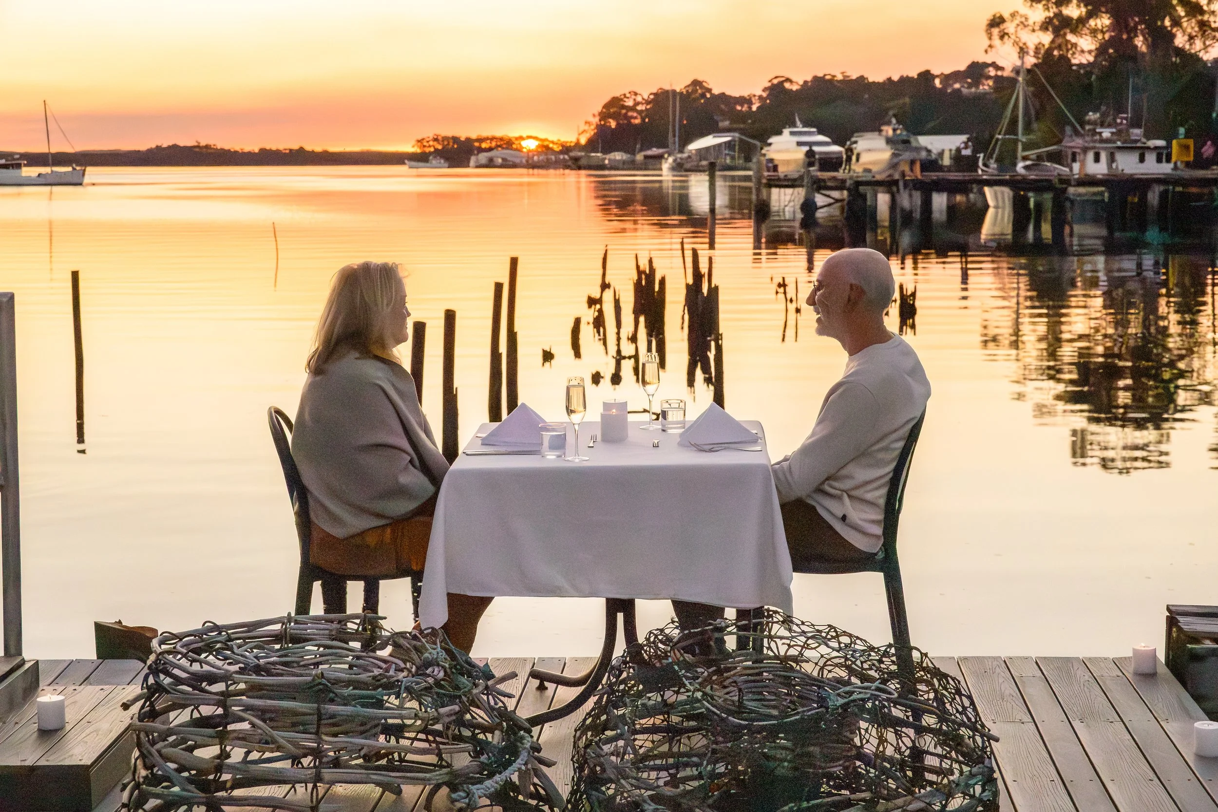 A couple dining at Risby Cove's Jetty Table by the water during sunset, with boats docked in the background and string lights overhead.
