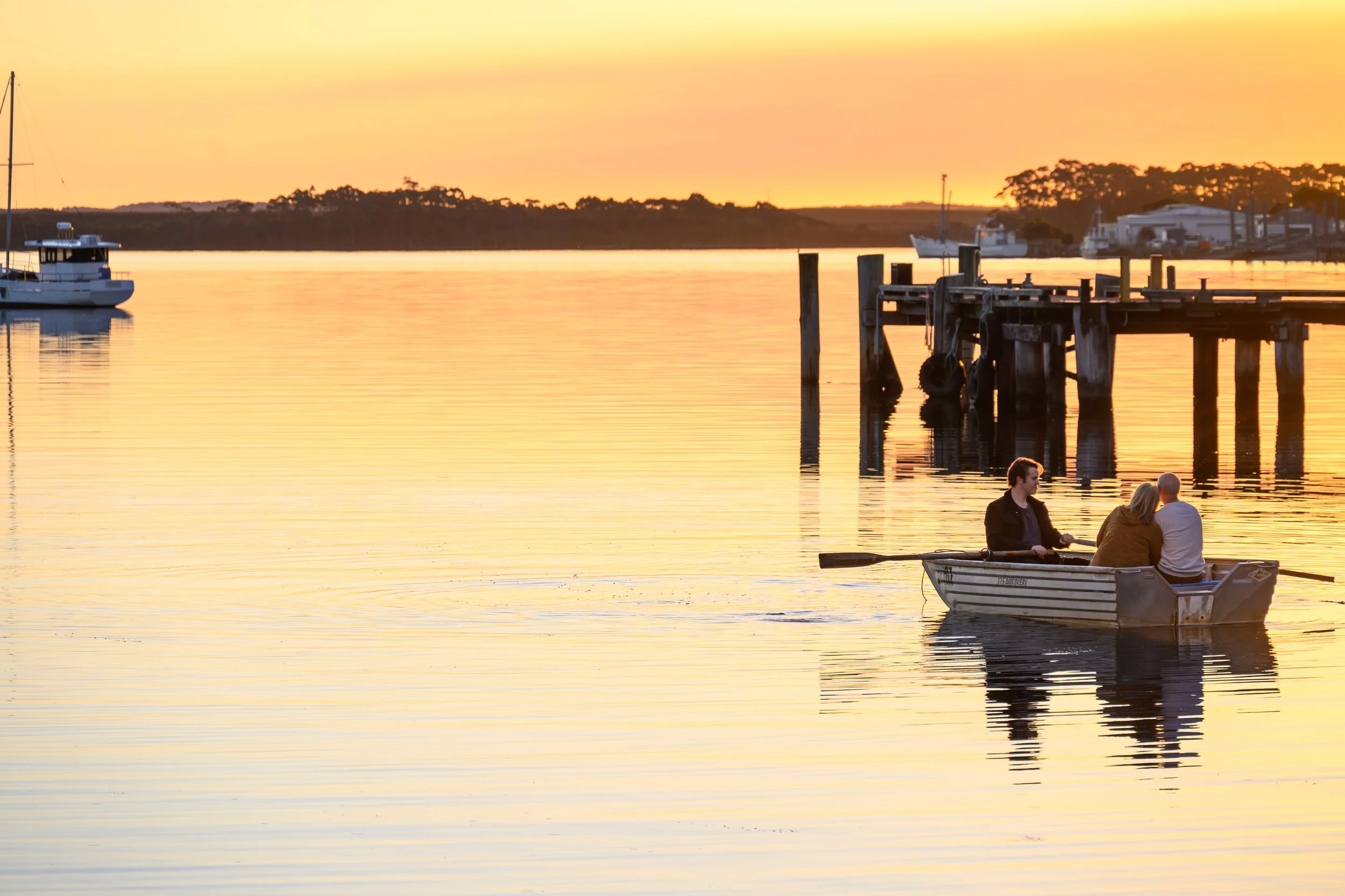 Three people in a small boat on a calm body of water during sunset, with boats and a wooden dock in the background.
