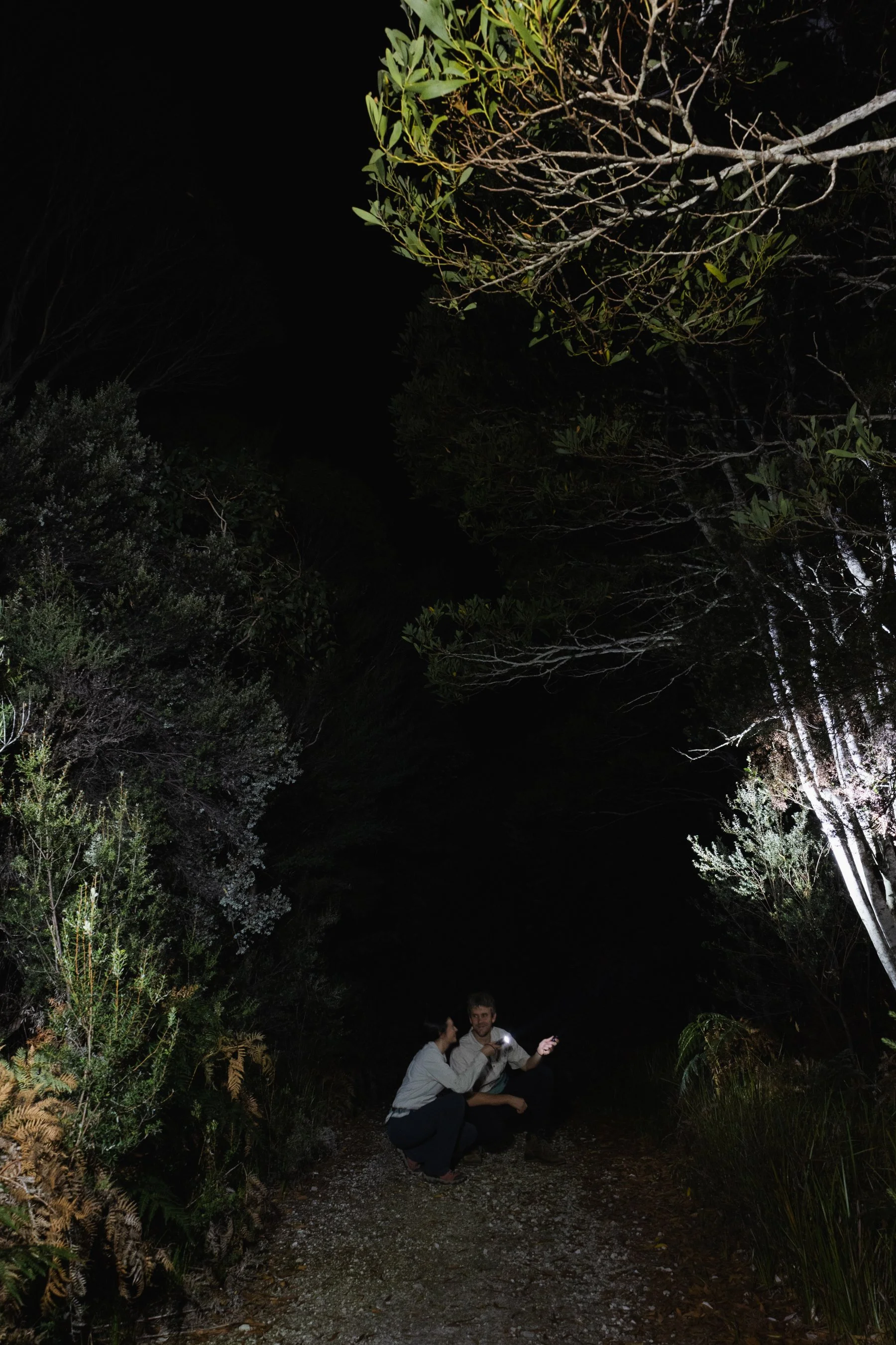 Two people squatting and smiling on a narrow dirt path during nighttime, surrounded by tall bushes and trees, with one of them holding a flashlight.