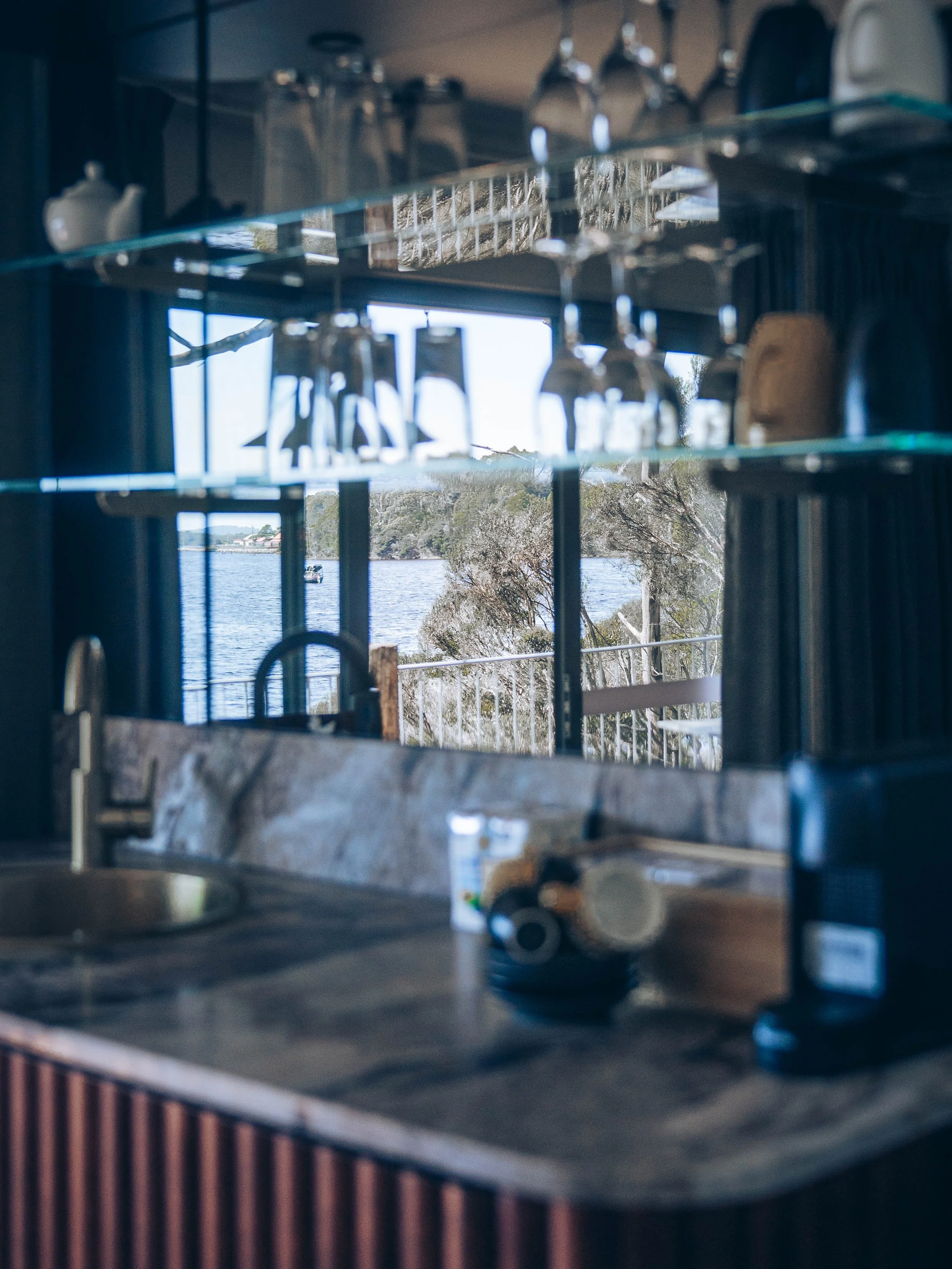 Bar unit counter with a sink, cups, and glasses, reflecting a view of Macquarrie Harbour and trees through a window.