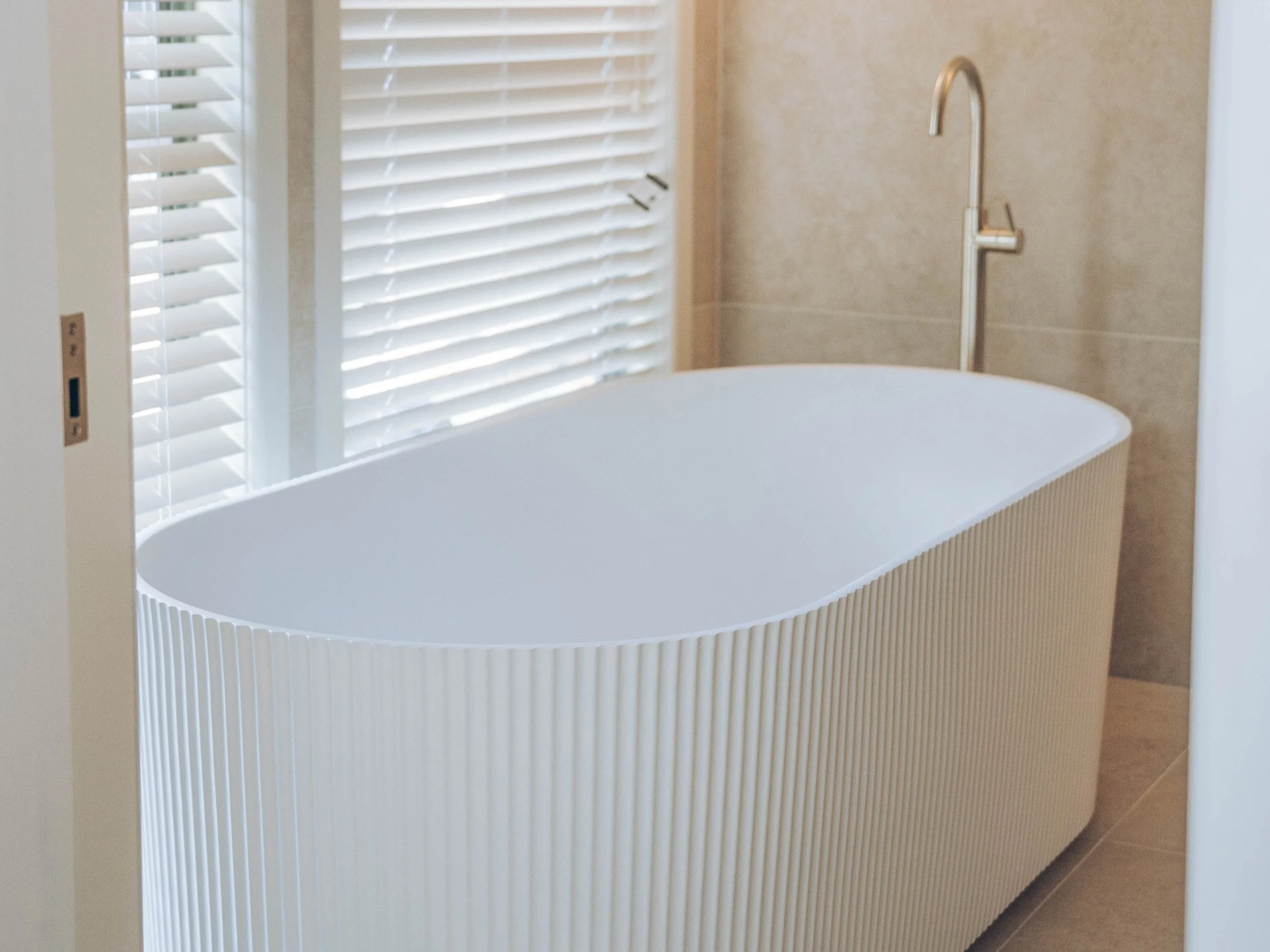Modern freestanding fluted bathtub and a sleek gold faucet, situated near a window with white blinds.