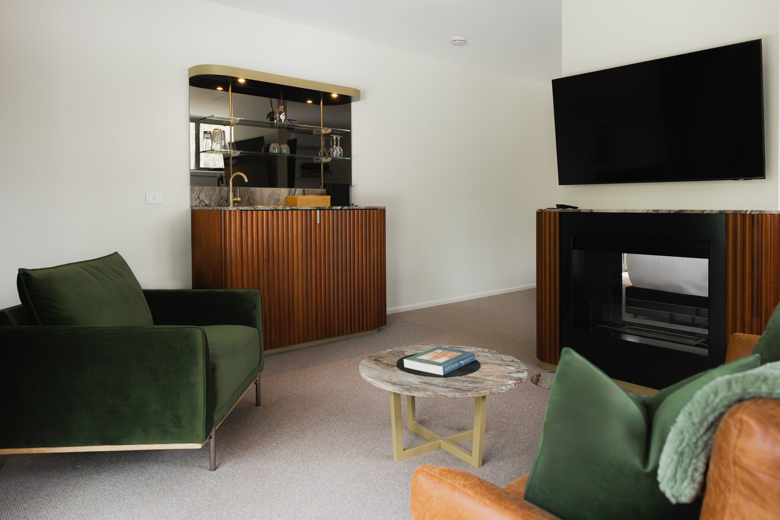 Living room with green velvet sofa, orange armchair, marble coffee table with books, wall-mounted TV, and a built-in fireplace with wooden paneling, in a modern style.