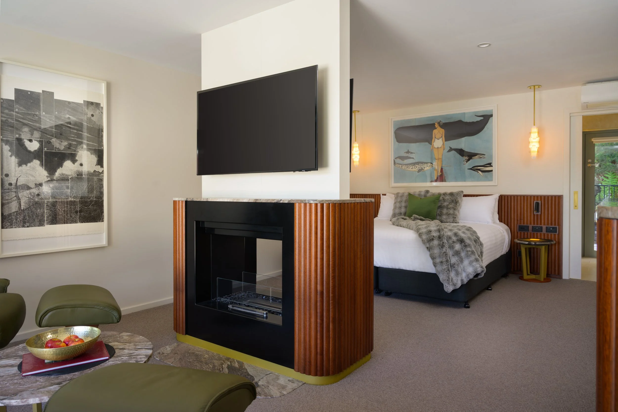 Modern hotel room with a king bed, artwork on the walls, wall-mounted television, fireplace, sitting area with green chairs, and a balcony door.