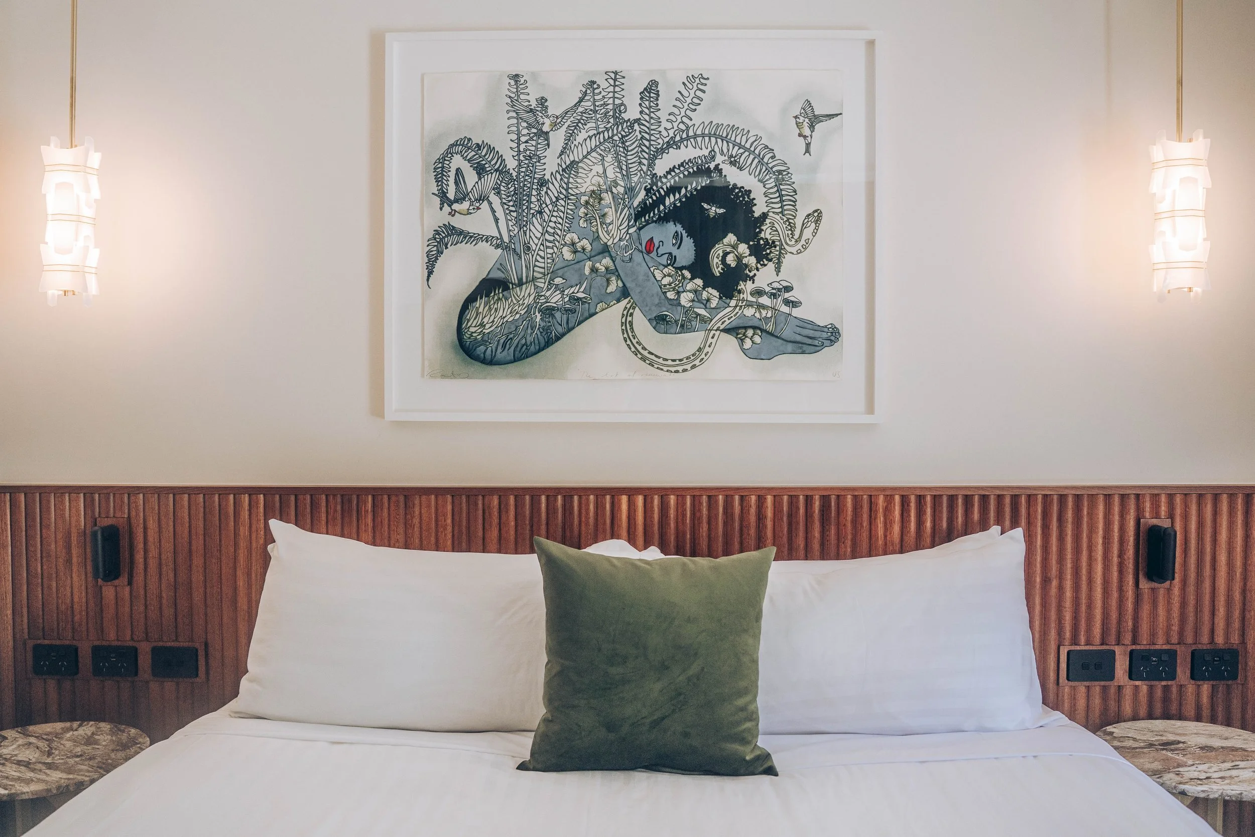 A bedroom with a bed, green pillow, white headboard, wall-mounted lamps, and a framed artwork of a sleeping woman surrounded by plants and animals.
