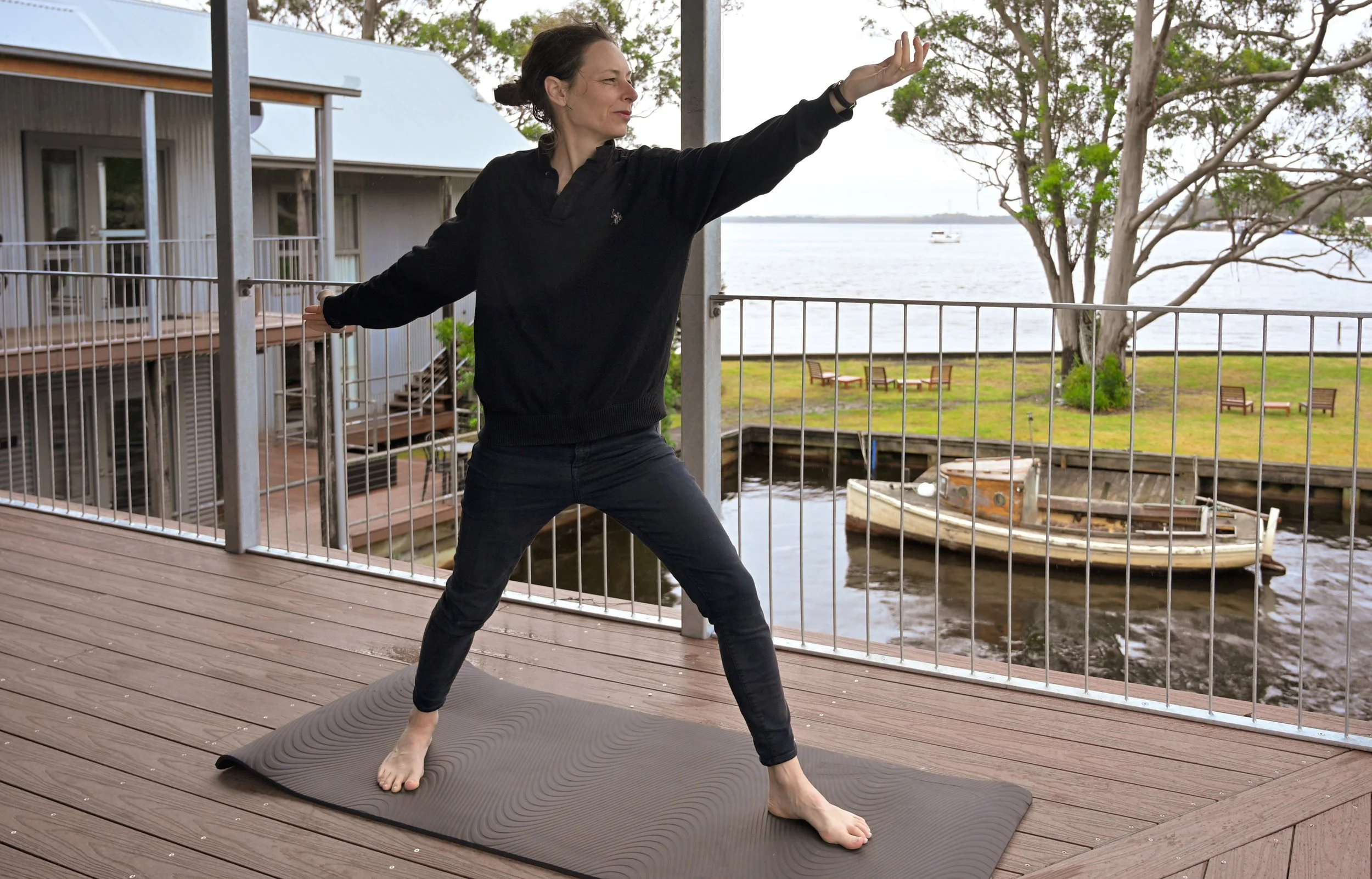 A woman practicing yoga outdoors on a wooden deck near the water, standing in a wide-legged stance with one arm raised and the other extended behind her, with trees and a boat in the background.