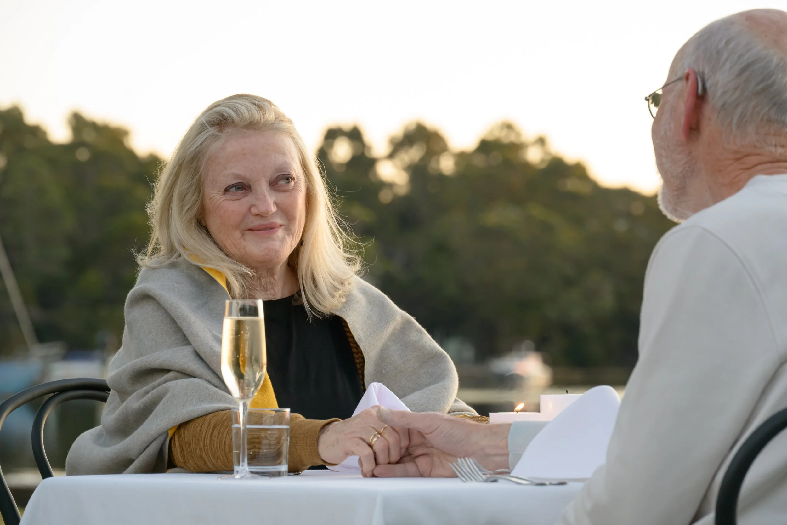 A woman with blonde hair, wearing a gray jacket, sitting at a dining table outdoors with a glass of champagne in front of her, holding hands with a man dressed in white, during sunset with trees in the background.