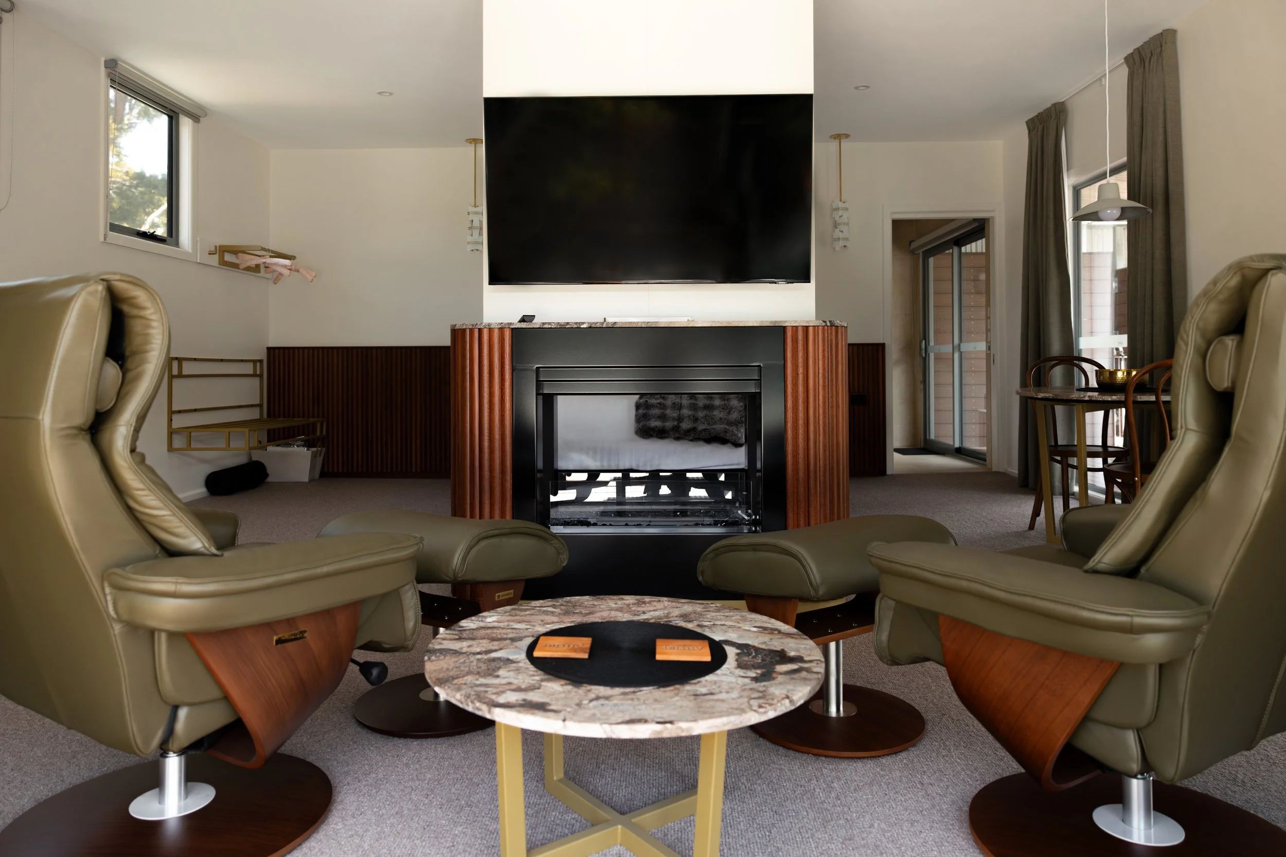 Living room with a fireplace, a large television, two green armchairs, a marble coffee table, and a dining area with a round table and four chairs.