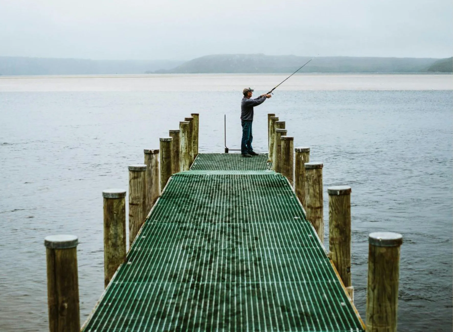 Man fishing on a green jetty at Macquarie Heads, extending into Macquarie Harbour with distant hills in the background