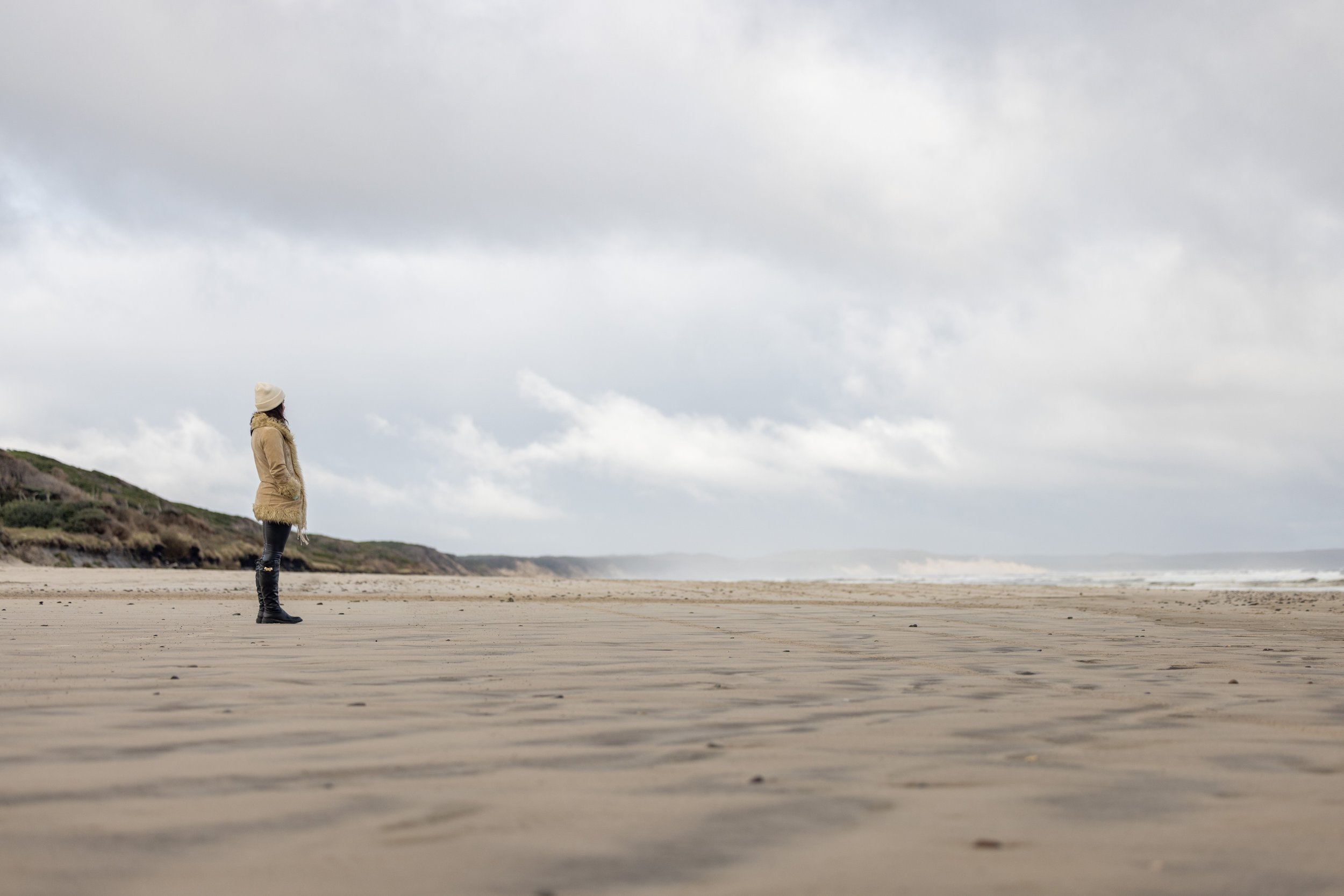 A person stands alone on Ocean Beach looking toward the ocean, wearing a beige coat, black pants, boots, and a white hat, under a cloudy sky.