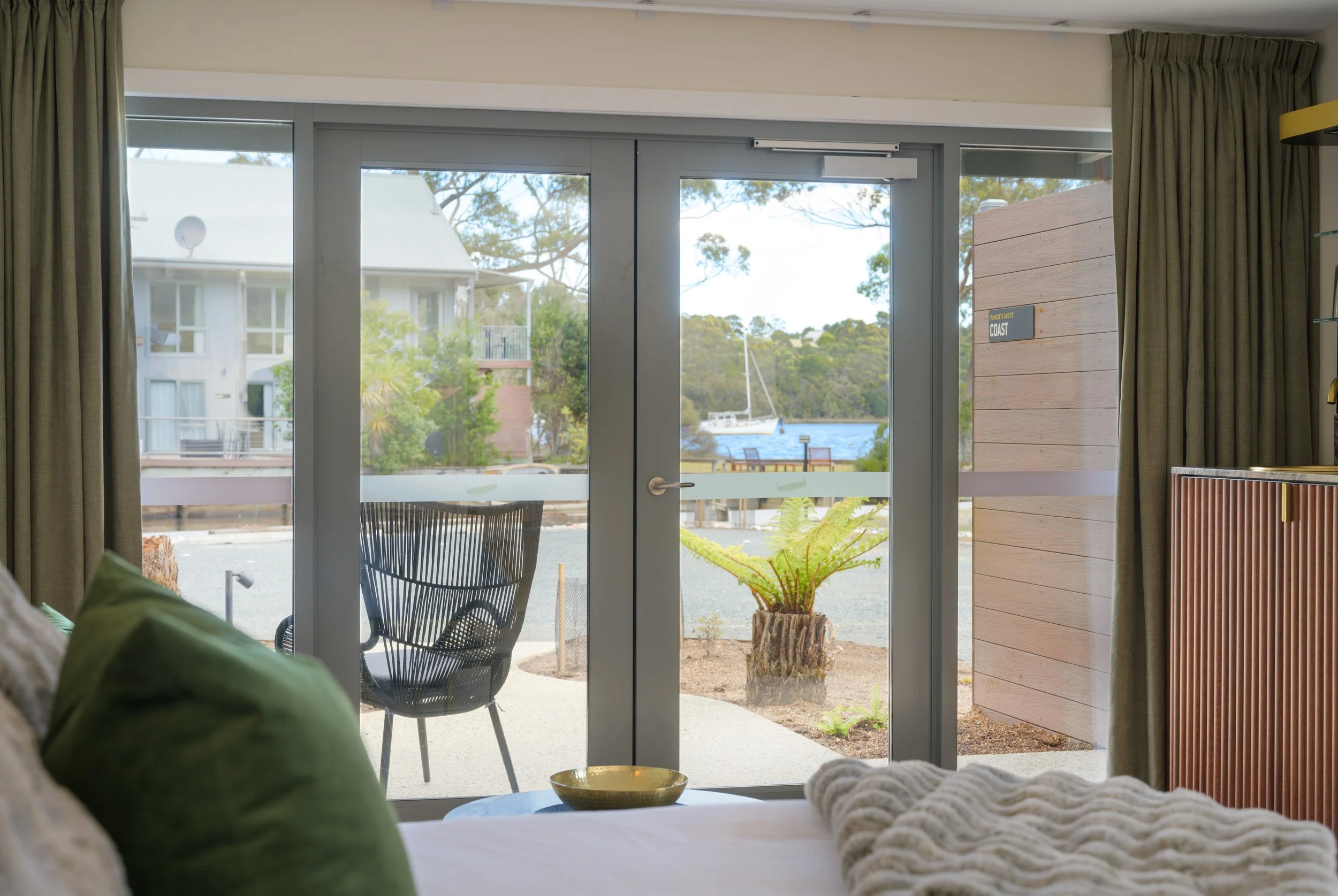 View of a glass door leading to an outdoor patio with a boat on the water, potted plants, and neighboring buildings in the background, seen from inside a room with green curtains and a bed with a green pillow.