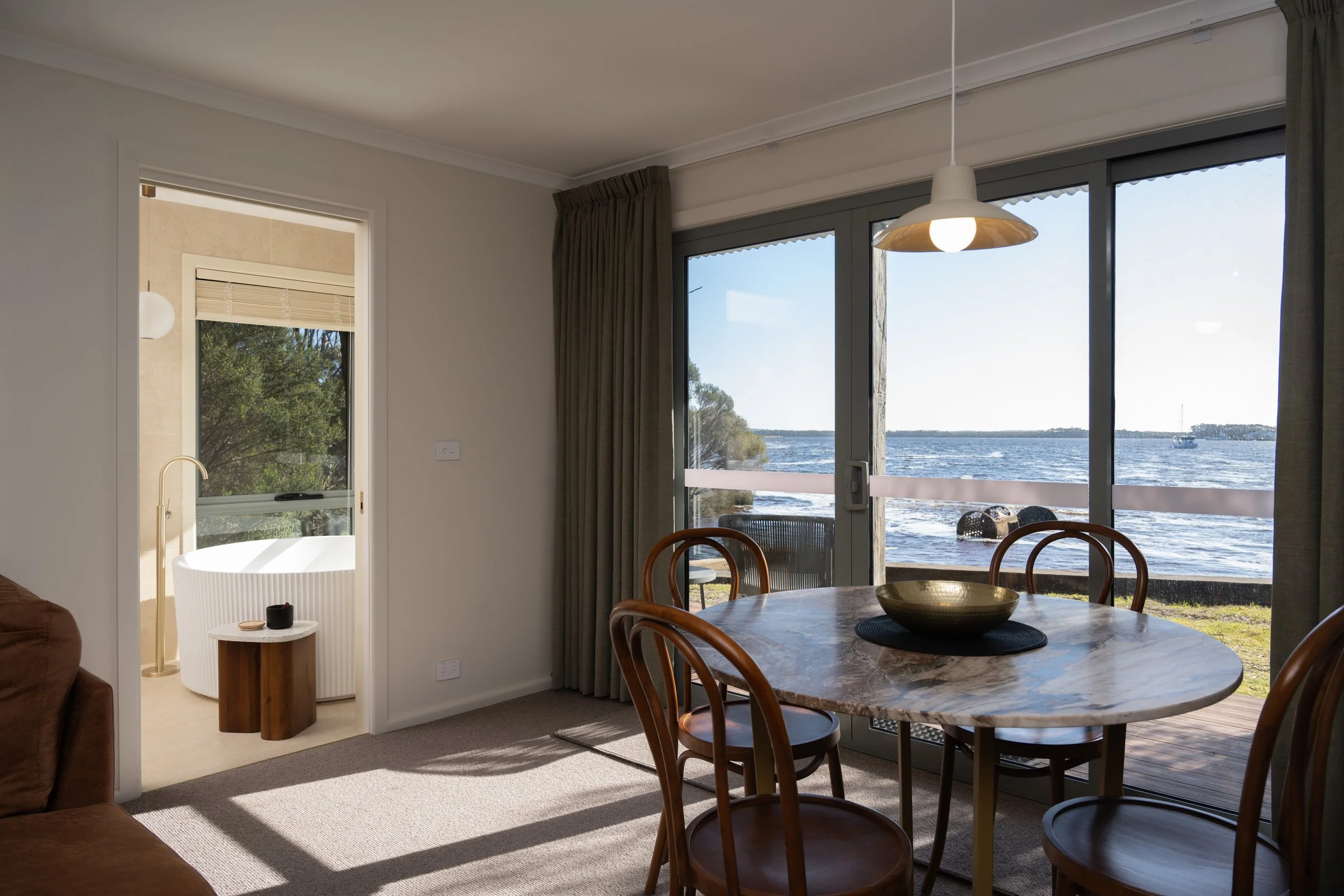 Living room with large glass doors opening to a river view, round dining table with four wooden chairs, curtains, pendant light, and a glimpse of a bathroom with a bathtub and a balcony outside.