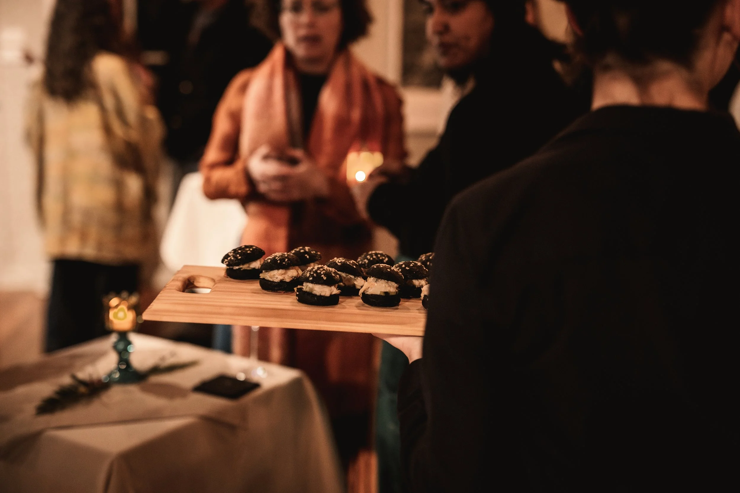A person holds a wooden tray with black lobster brioche canapés, with several other people in the background at a gathering or party.