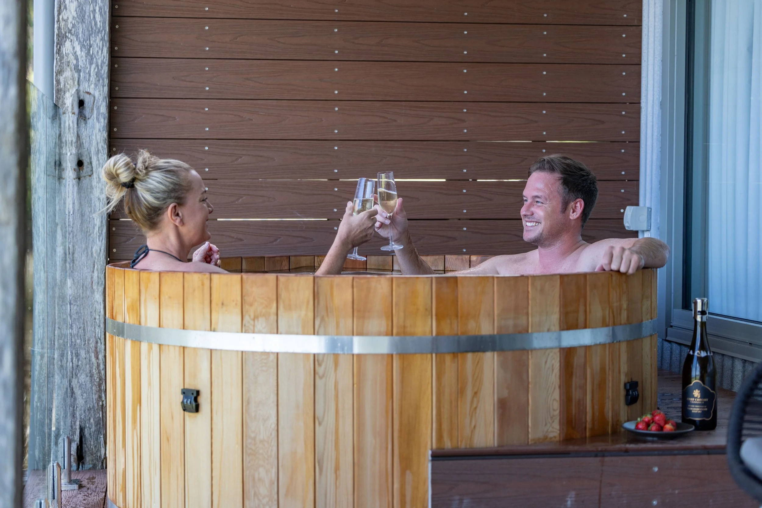 A man and woman relaxing in a wooden hot tub on a balcony, sipping champagne and smiling at each other, with a bottle of champagne and bowls of strawberries nearby.