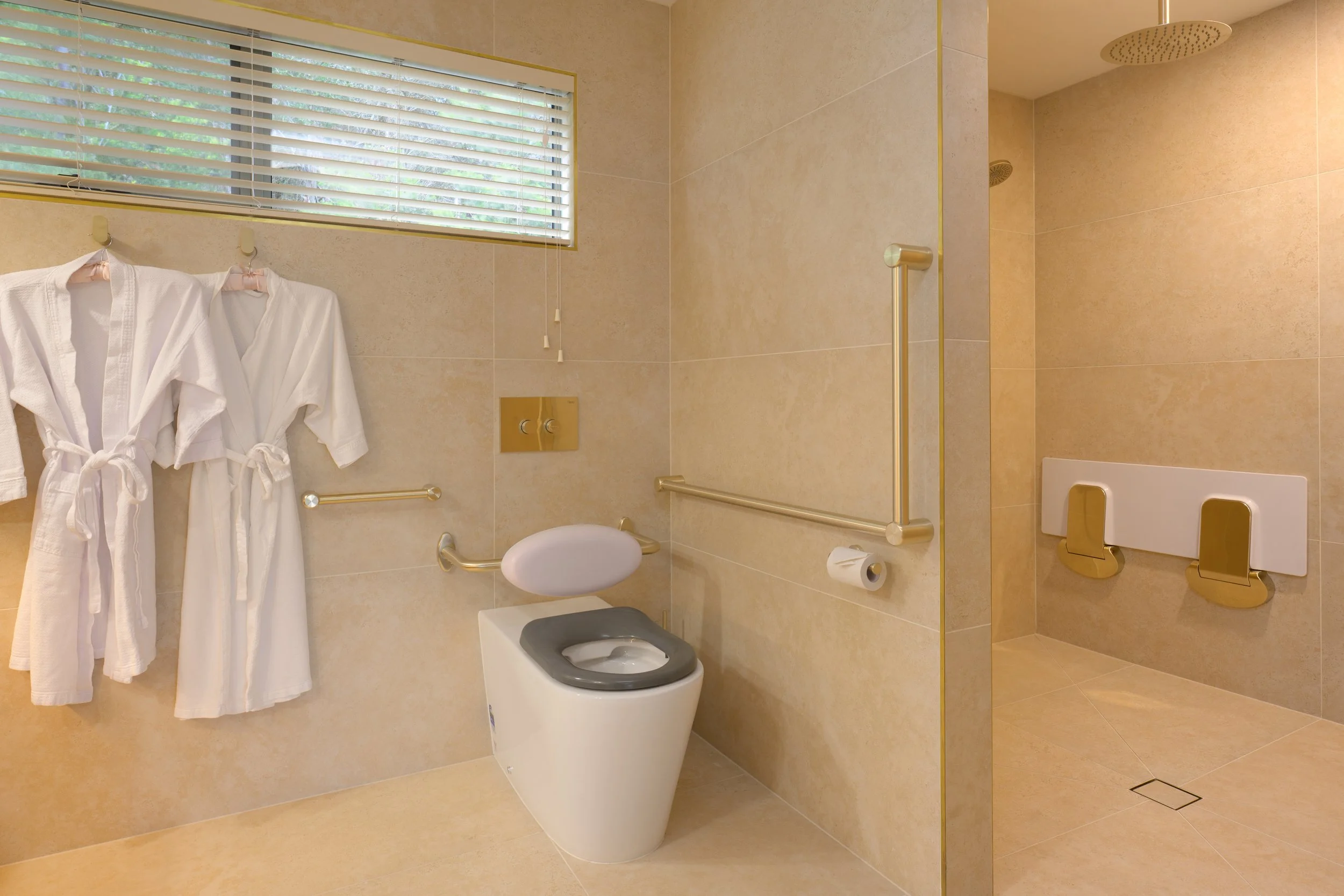 Accessible bathroom with a toilet, grab bars, two white bathrobes hanging on a rail, a window with horizontal blinds, and a shower area with additional grab bars.