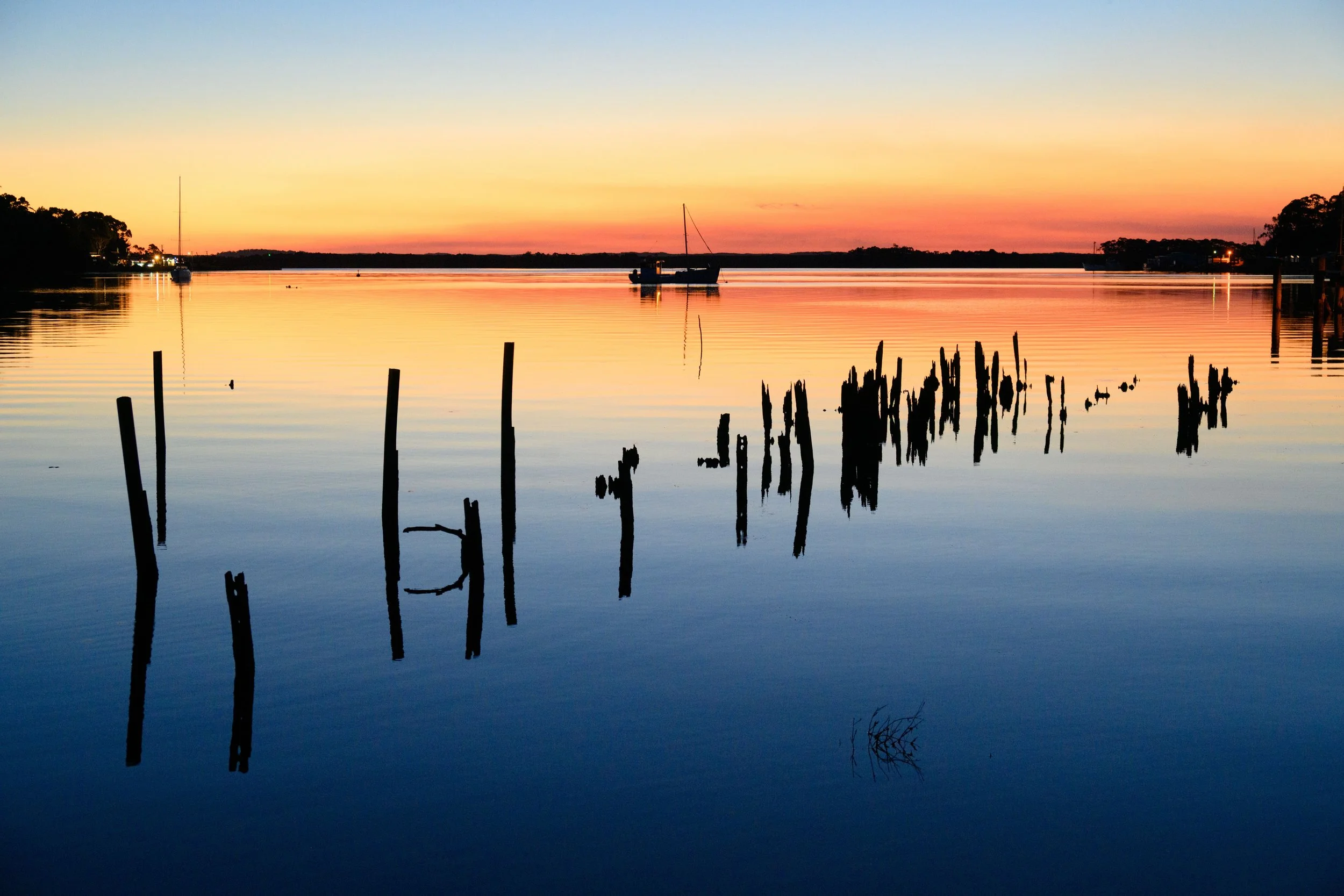 Sunset over Macquarie Harbour with boats and silhouetted trees, ruins of an old jetty in the water.