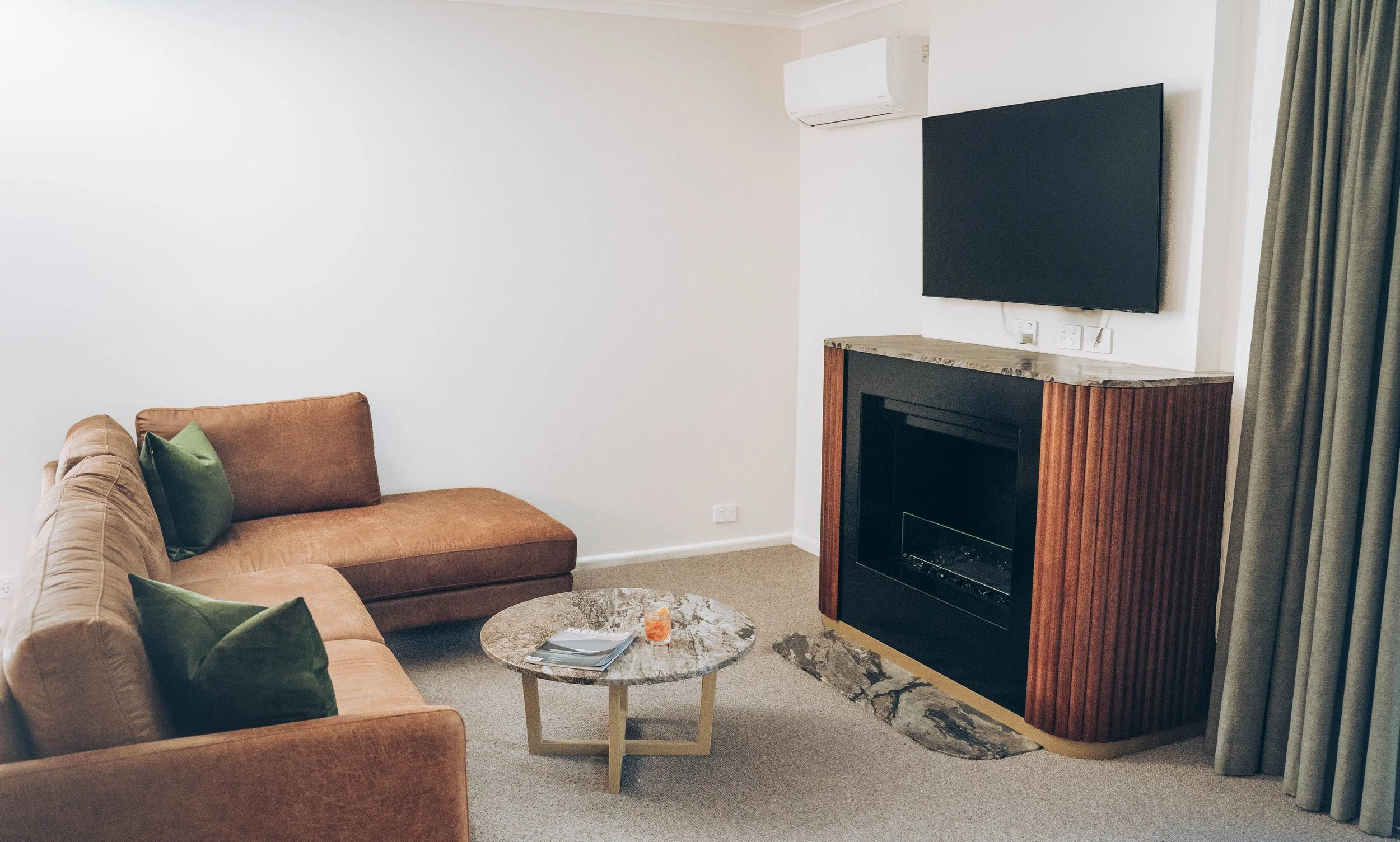 Living room with a tan sofa, green pillows, a round marble coffee table with magazines and a decorative candle, a fireplace with a marble top, a wall-mounted TV, an air conditioning unit, and a beige curtain.