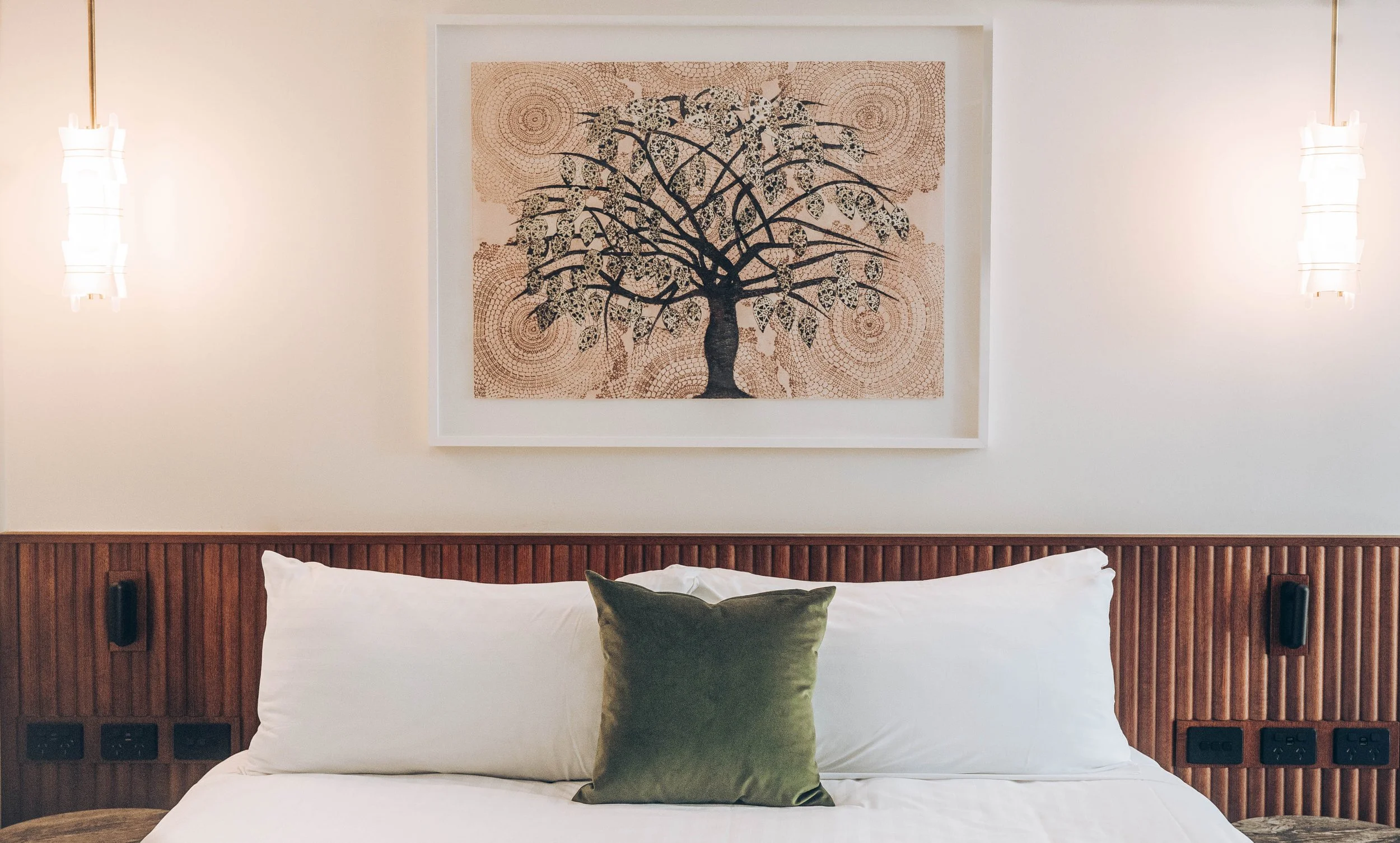 A neatly made bed with white linens and a green pillow against a wooden headboard. Above the bed is a framed artwork featuring a stylized tree with patterned leaves, flanked by two modern wall sconces.