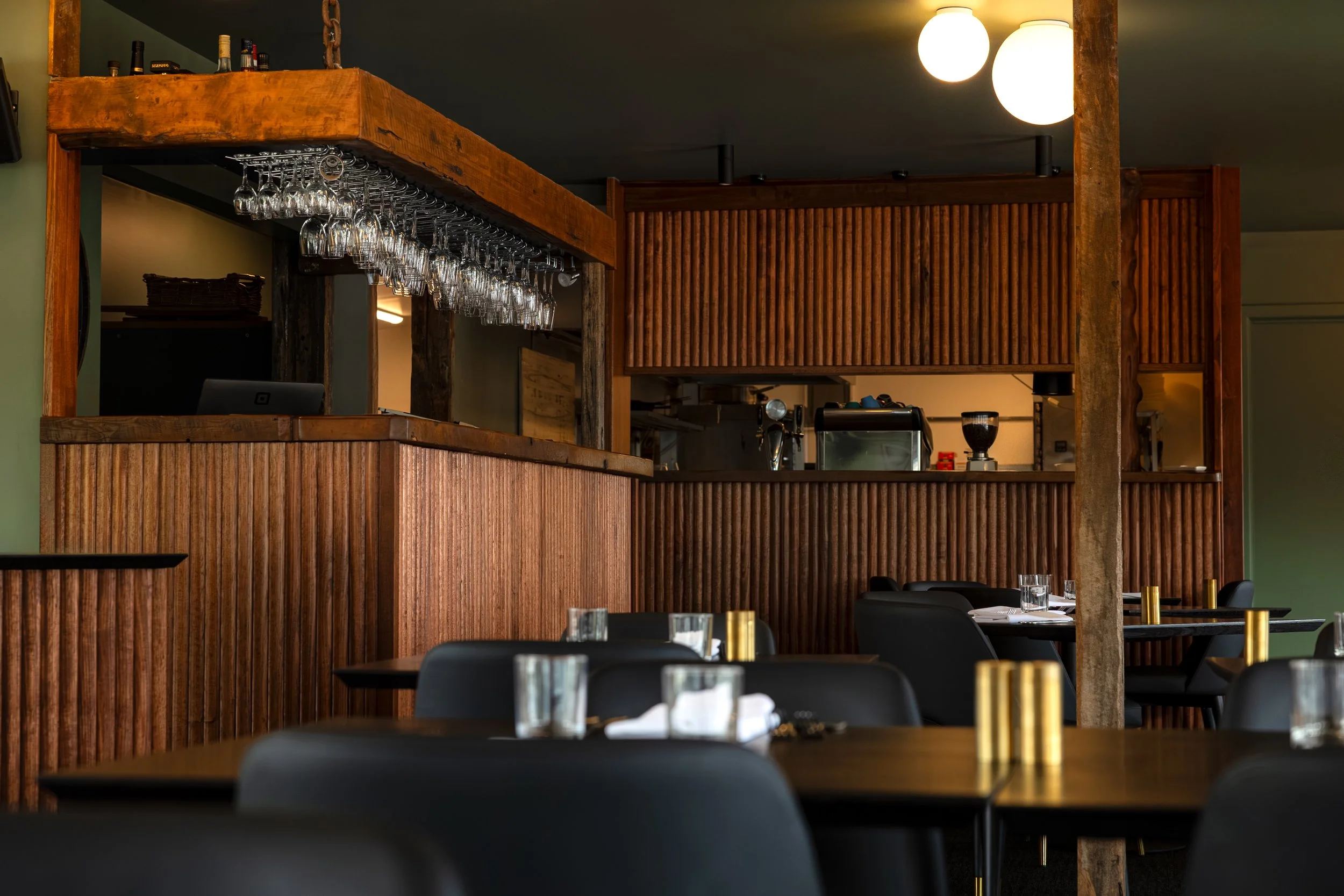 Interior of a restaurant with wooden decor, black chairs, tables set with glasses and napkins, and a counter with hanging glassware and kitchen equipment.
