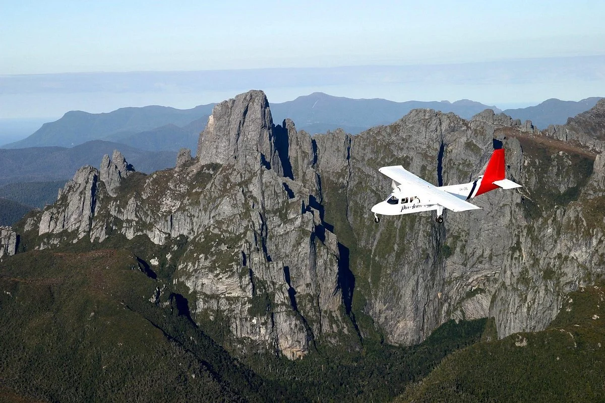 Small white airplane with red and black tail flying near rocky mountain cliffs over the Tasmanian Wilderness World Heritage Area.