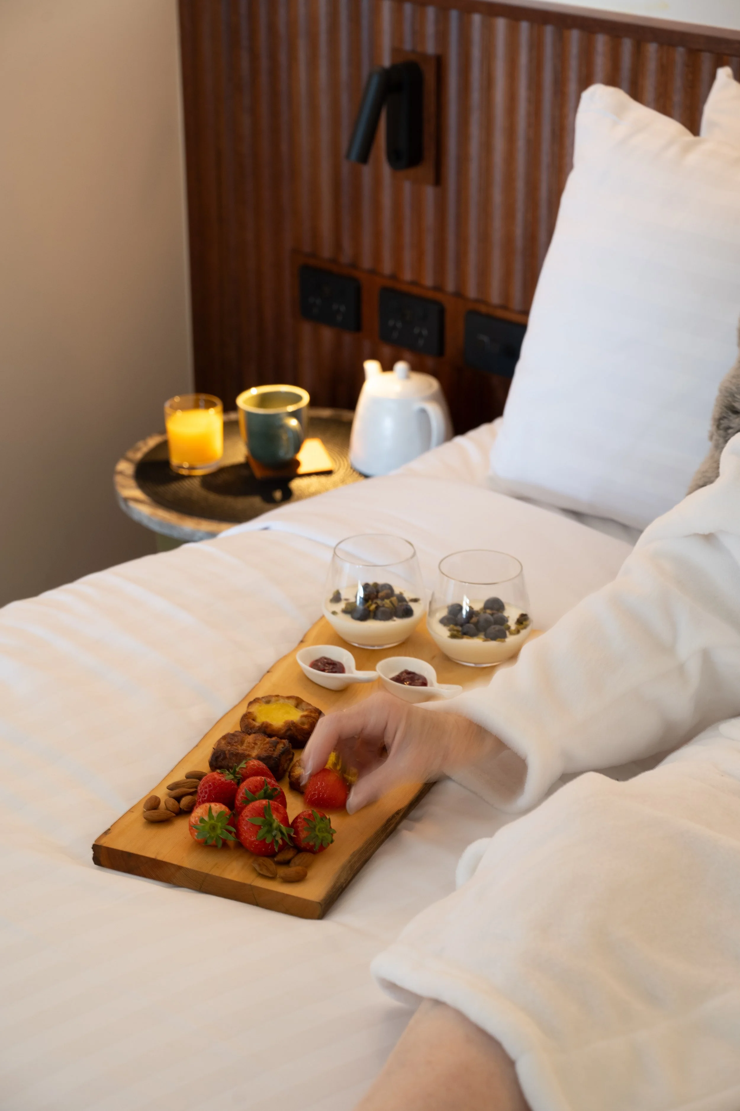A person is serving breakfast on a wooden tray on a bed, with strawberries, almonds, muffins, and small bowls of jam. There are two glasses of yogurt with blueberries behind, a cup, and a teapot on a nightstand. The bed has white sheets and pillows, and the room has wooden accents and multiple electrical outlets.