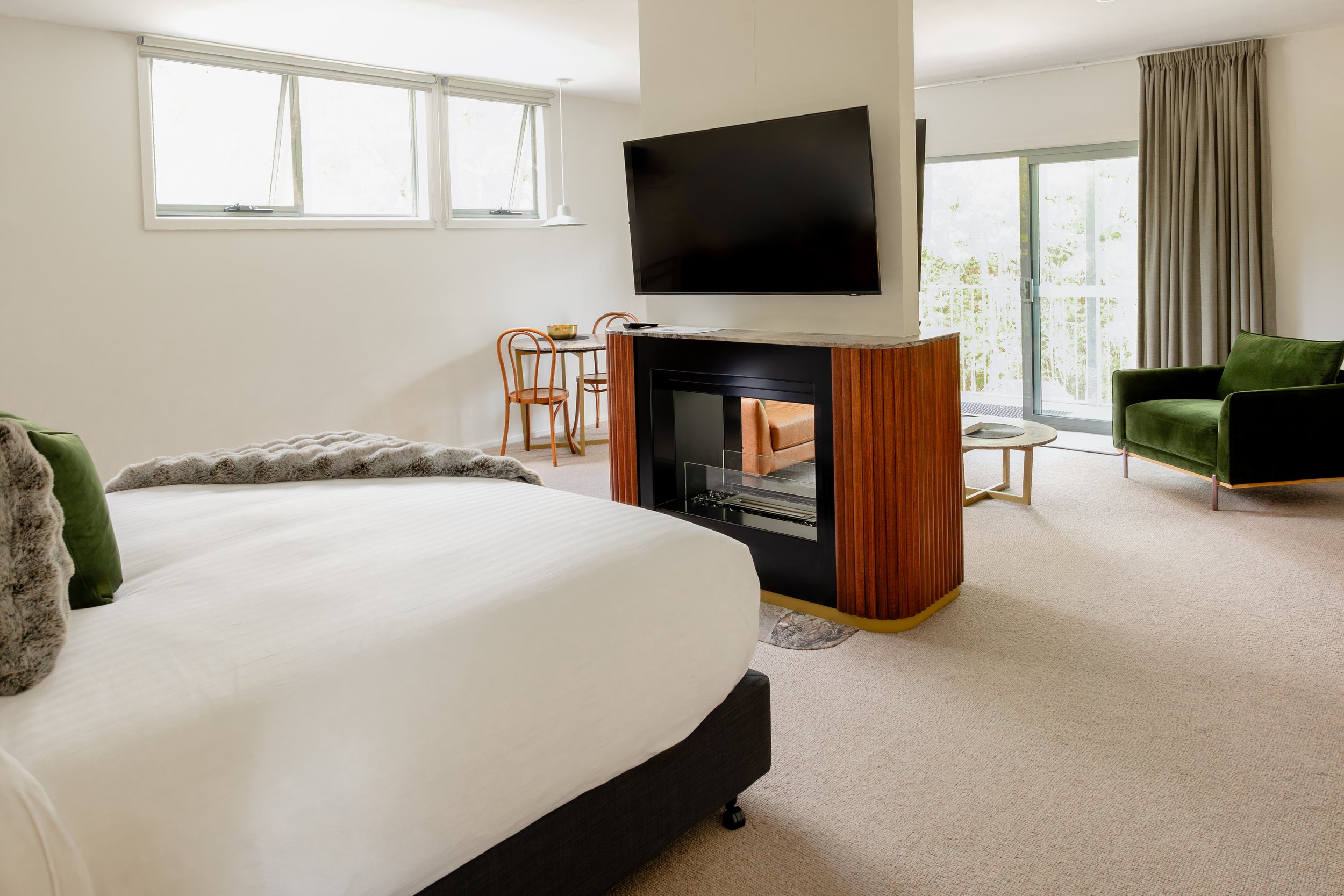Modern hotel room with a large bed, green armchair, fireplace, TV, sitting area with small dining table and chairs, and sliding glass doors leading to a balcony.