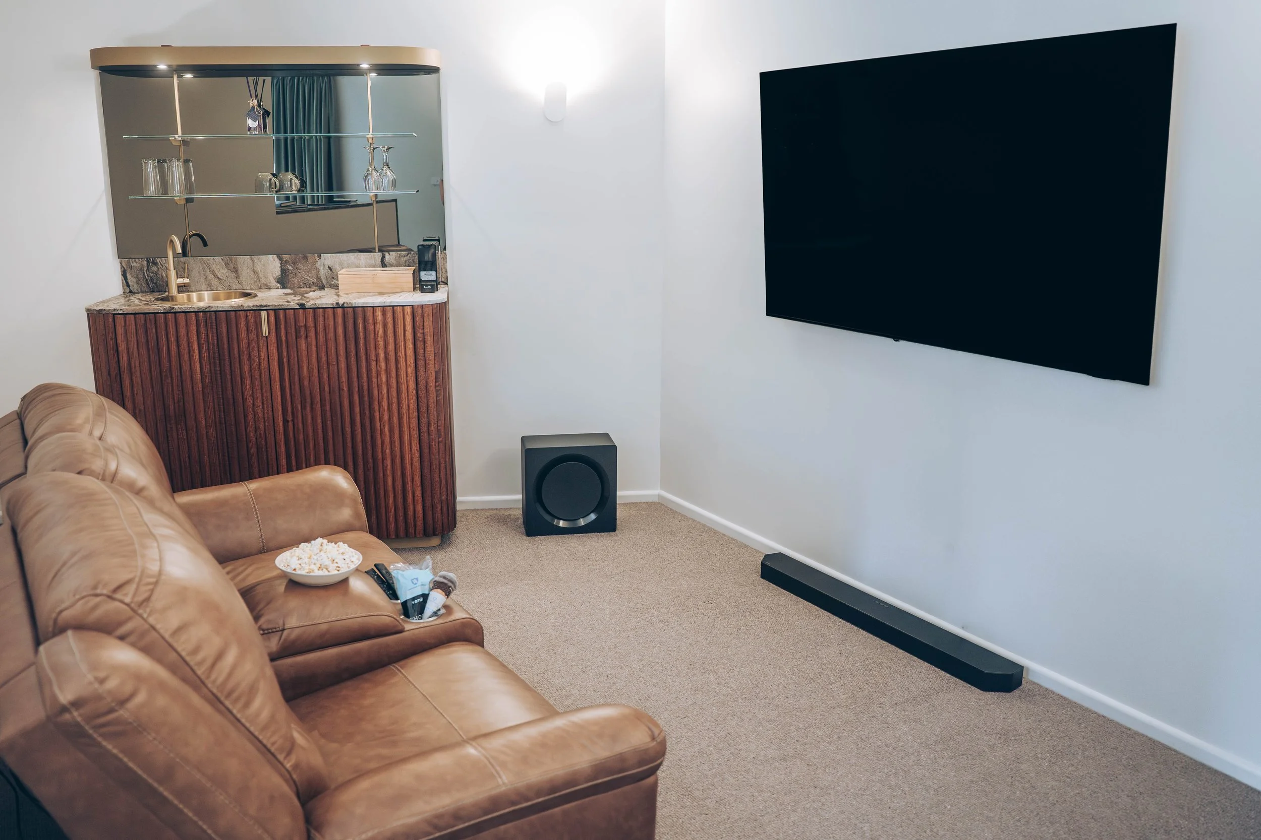 Theater living area with reclining leather sofas, a large smart TV, sound system, bar unit with sink and glass shelves, and a bowl of popcorn and pop-top ice cream on the armrest.