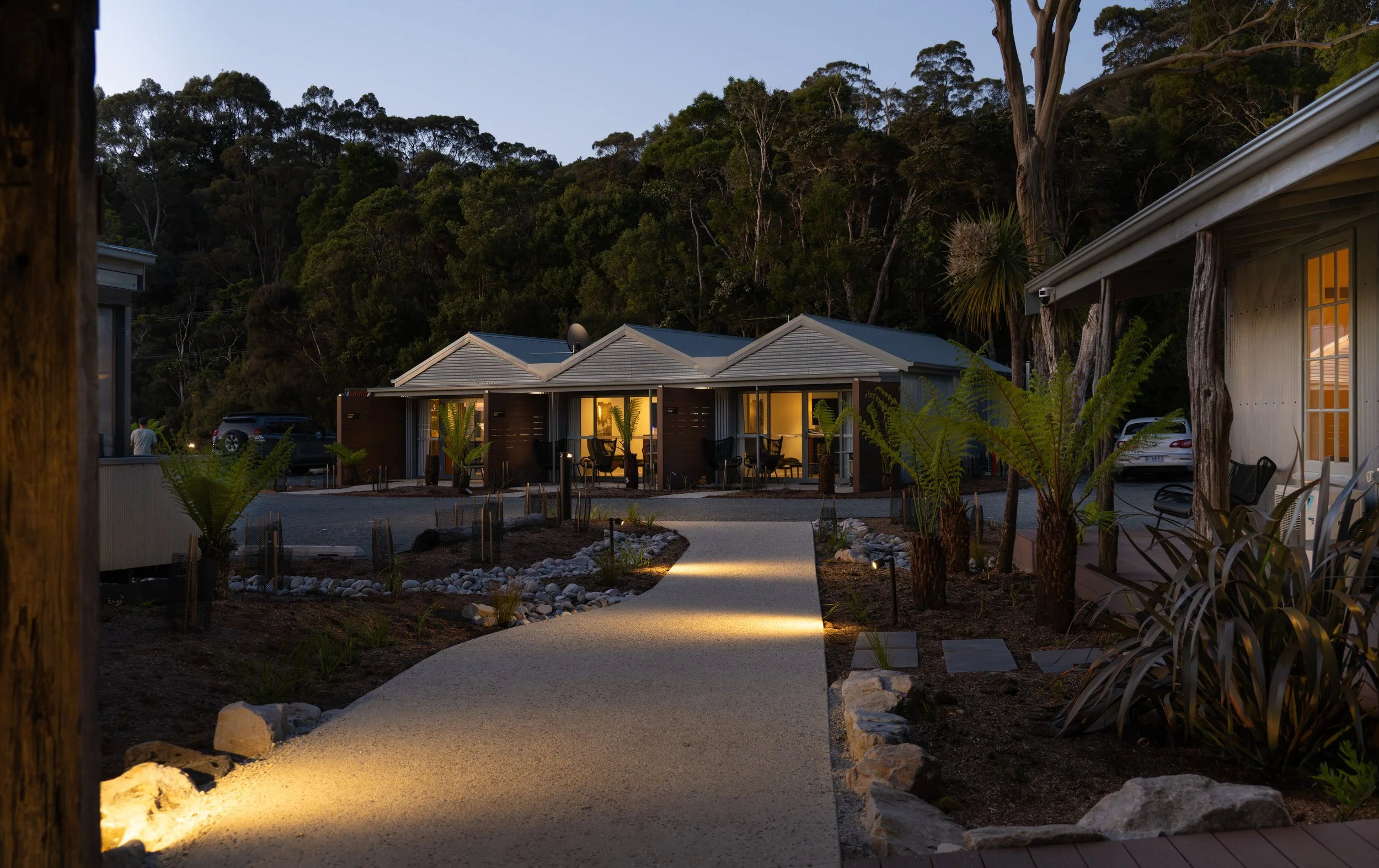 A pathway illuminated with ground lights leads to a row of suites with soft, warm lights on, set against a backdrop of a wooded hillside at dusk.