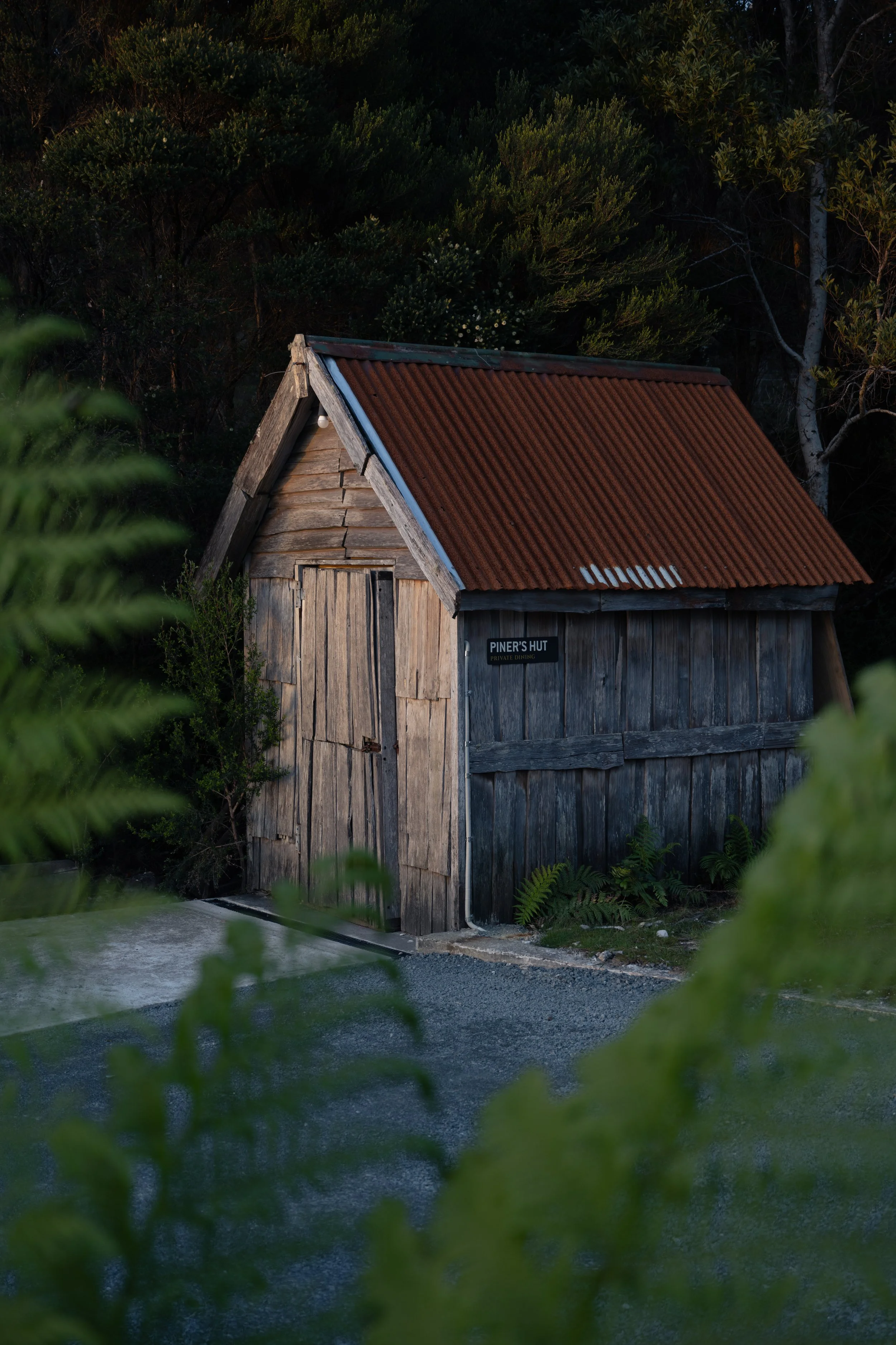 A rustic wooden hut with a rusted metal roof, surrounded by greenery and trees, with a sign that reads 'Piner's Hut Private Dining'.