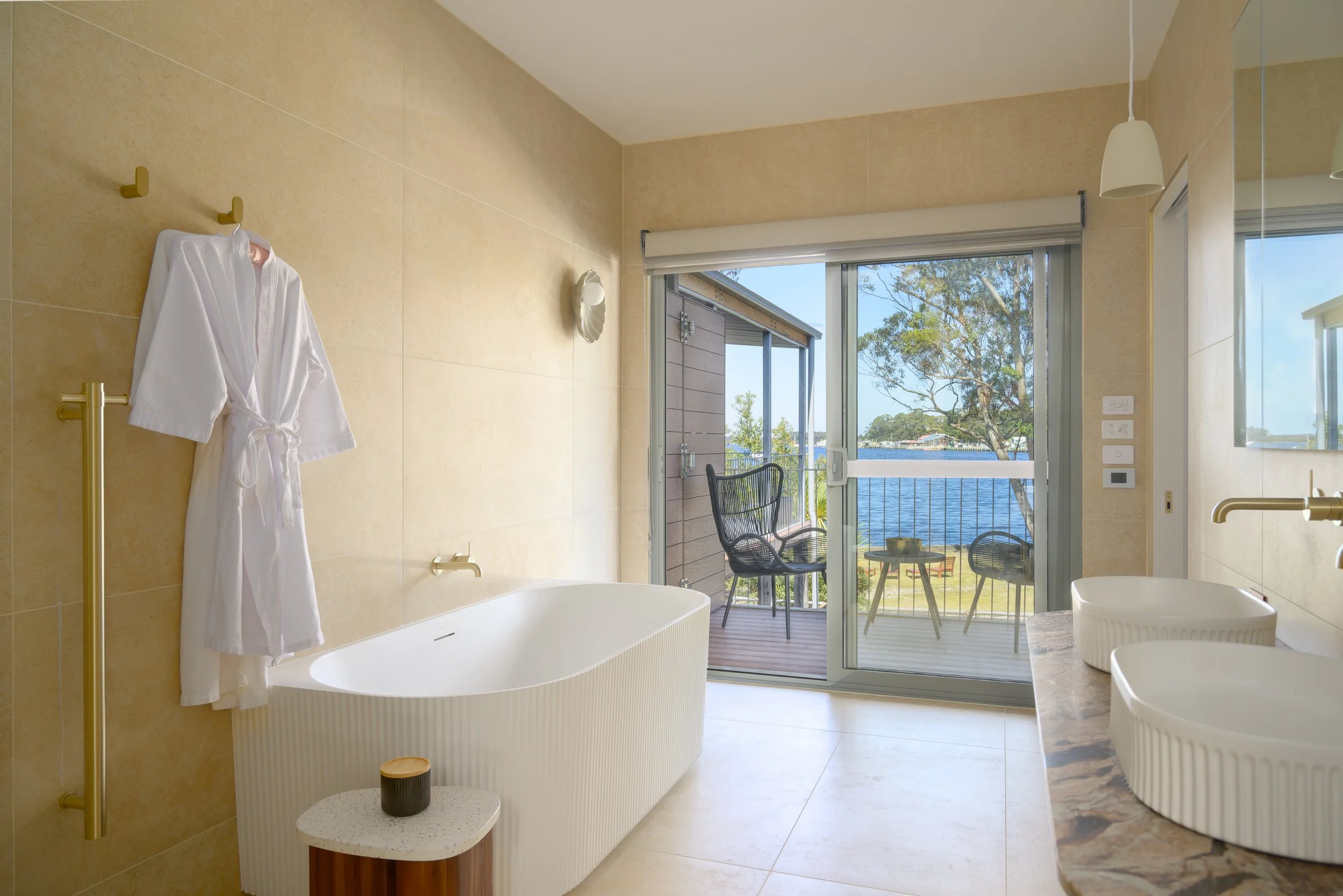 Bathroom with view of water and trees through sliding glass door, contains bathtub, double sinks, and a robe hanging on a gold towel rack.