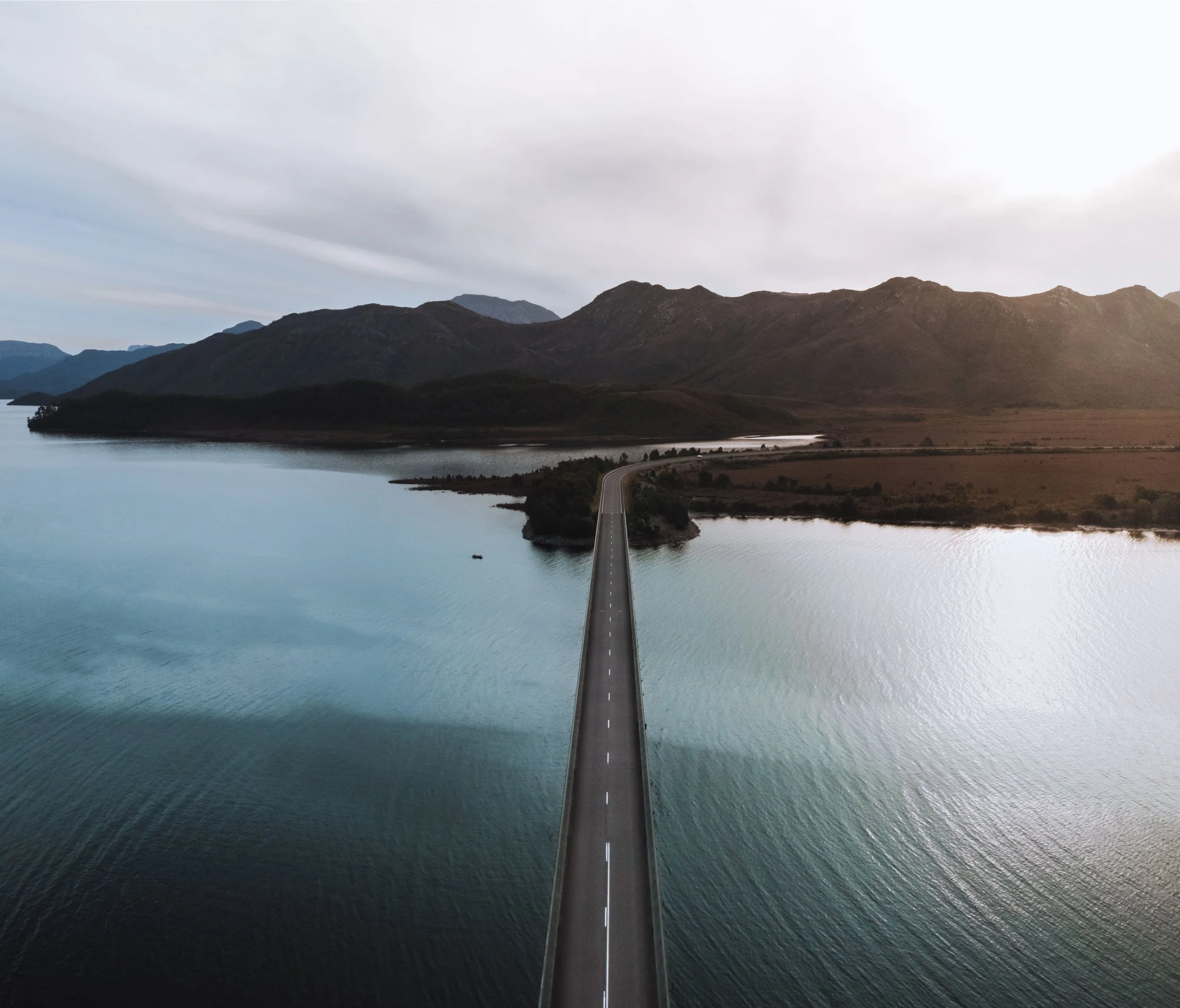 Aerial view of Bradshaw bridge crossing a Lake Burbury, a glacial lake with mountains in the background, on a cloudy day.