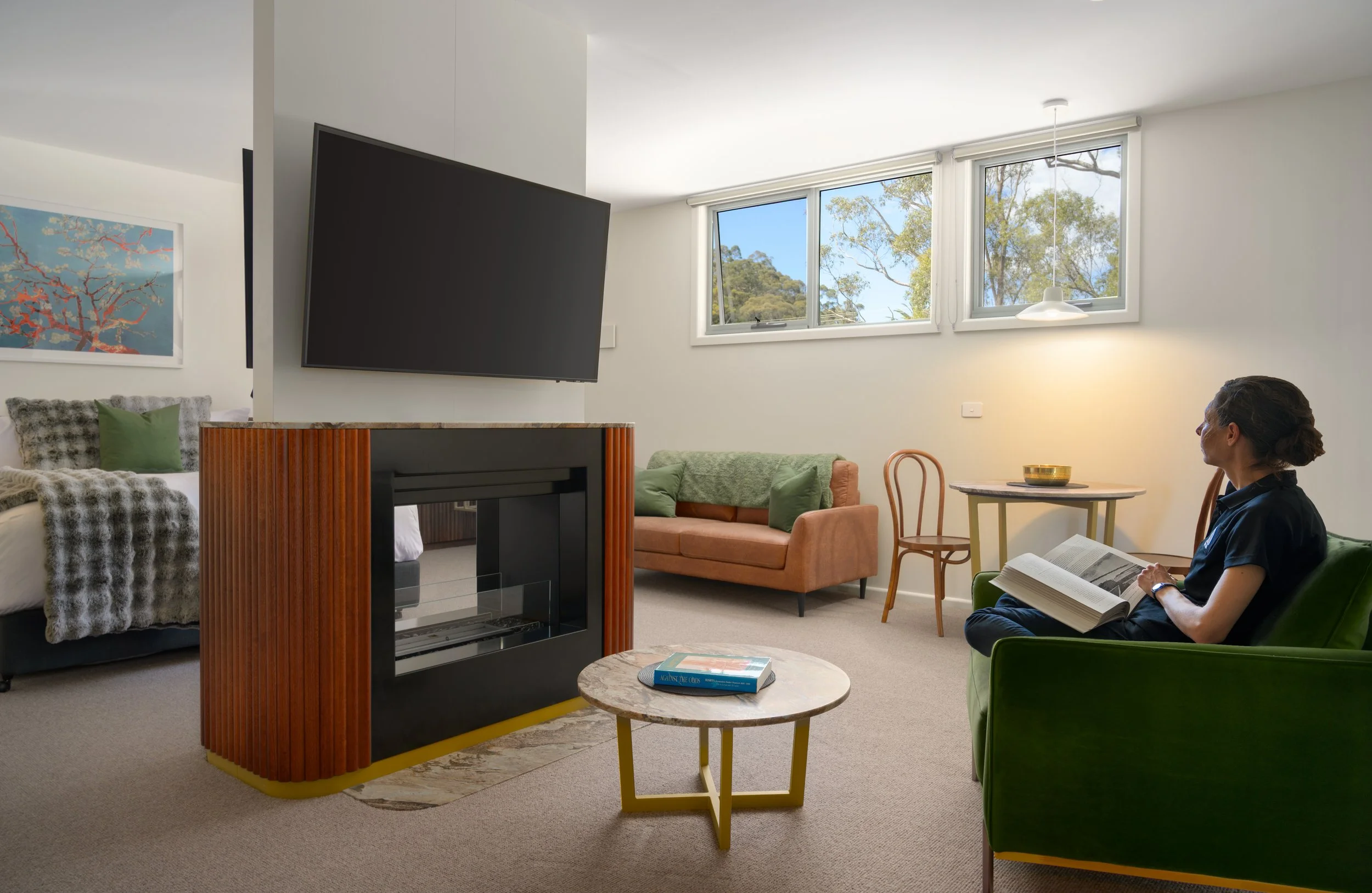 Living room with woman reading a book, green and peach sofas, wall-mounted TV, fireplace divider, small round coffee table, and windows showing trees outside.