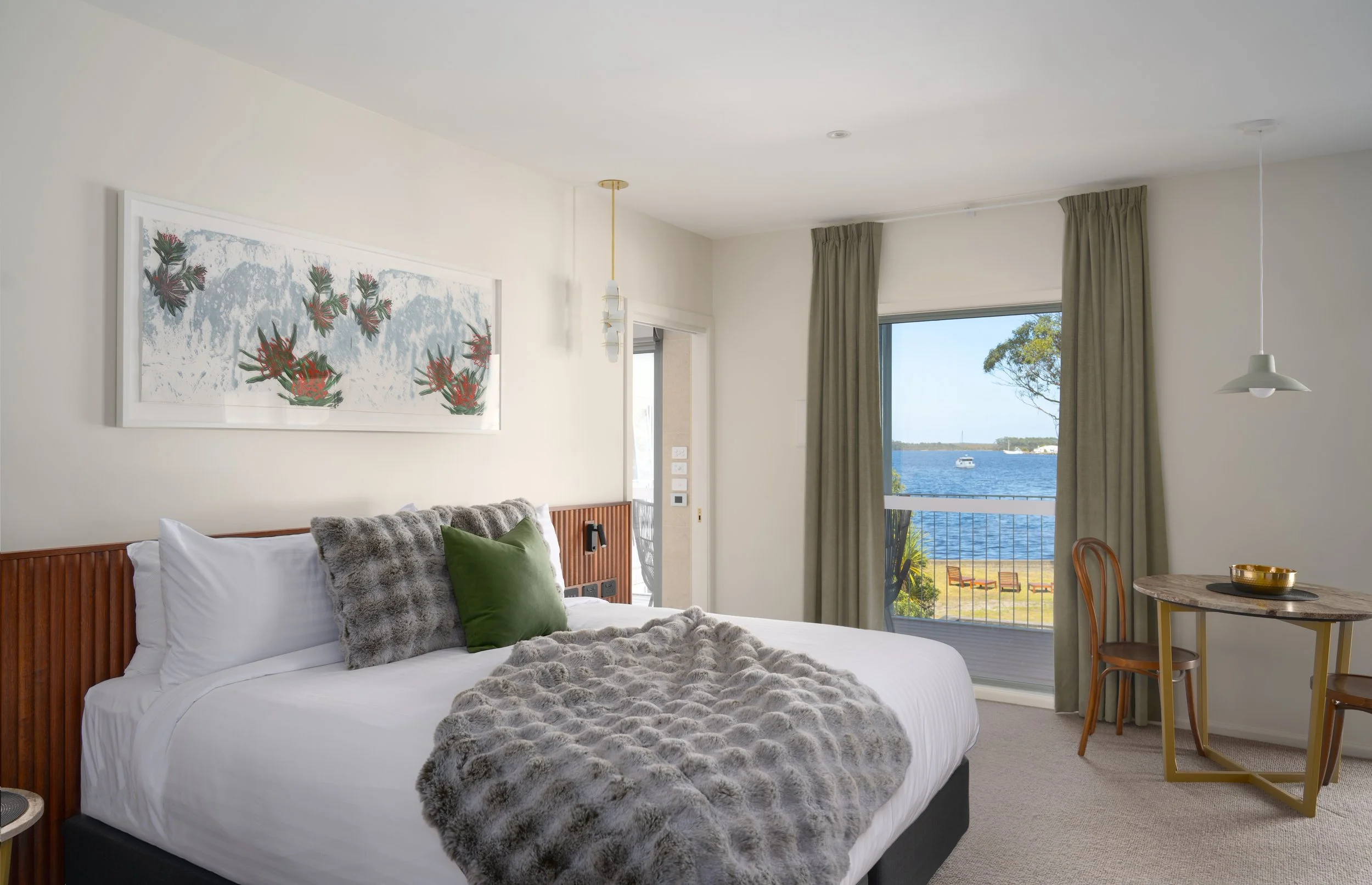 A bedroom with a large bed covered in white sheets, a grey faux fur throw, and pillows, one green and one grey. There is a window with green curtains showing a view of water, a boat, and trees outside. A round table with two wooden chairs sits near the window. A hanging pendant light and a wall-mounted art piece with floral designs decorate the room.