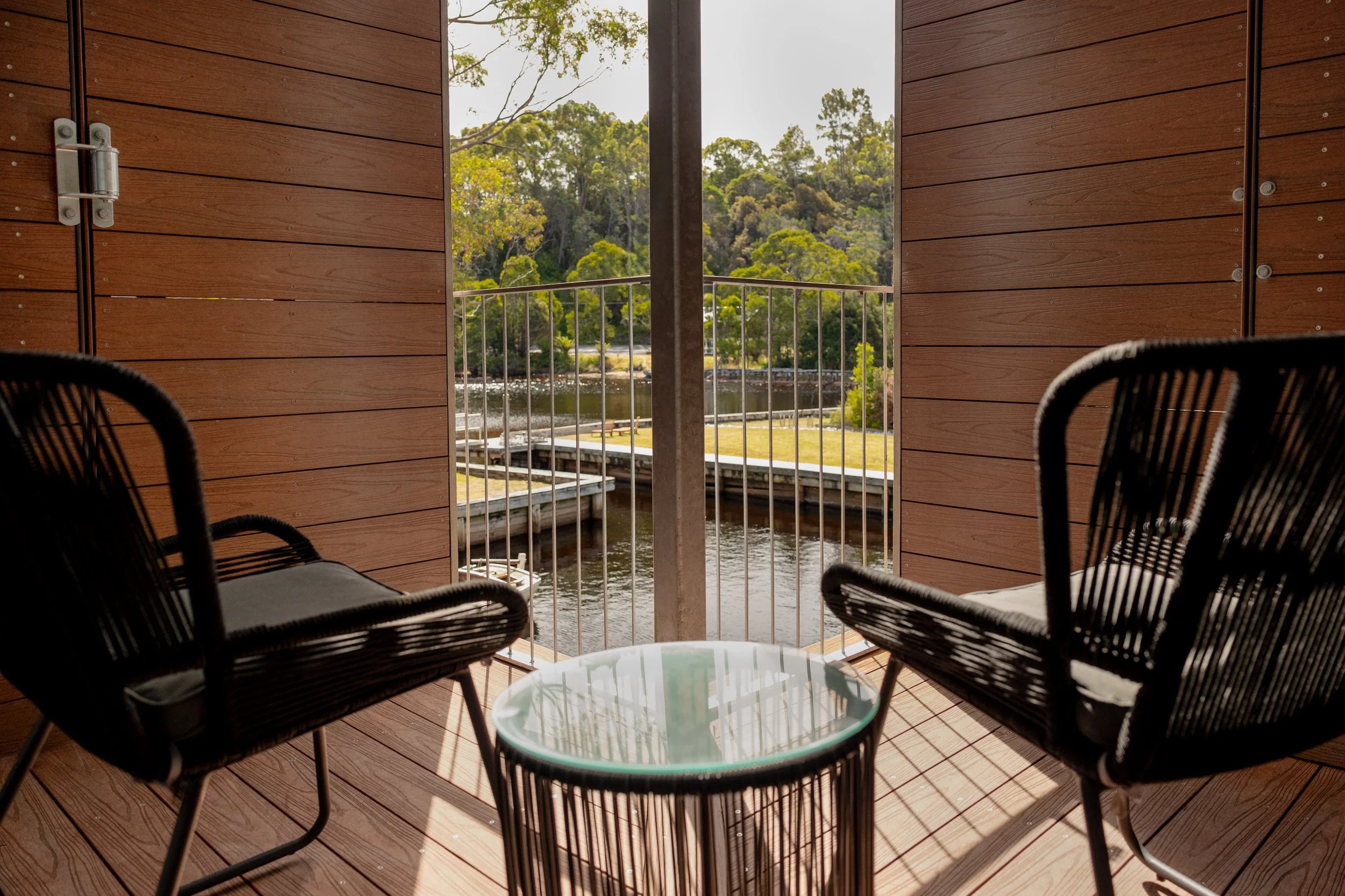 View from a wooden balcony with two black chairs and a small glass-topped table, overlooking a waterway with trees and greenery in the background.