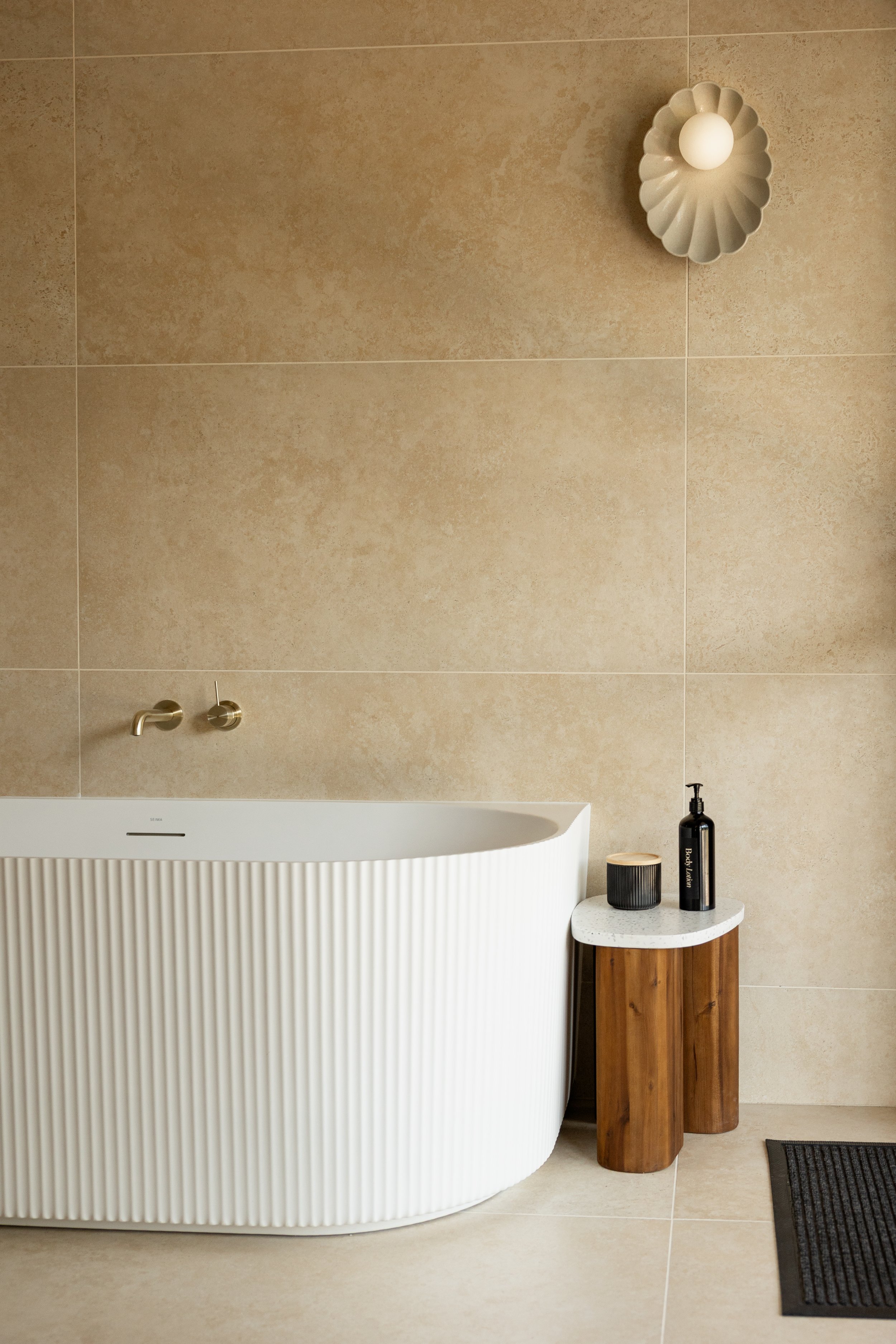 Modern bathroom with a white textured bathtub, beige tiled wall, small side table with black and natural decor, black soap dispenser, and a wall-mounted lamp.