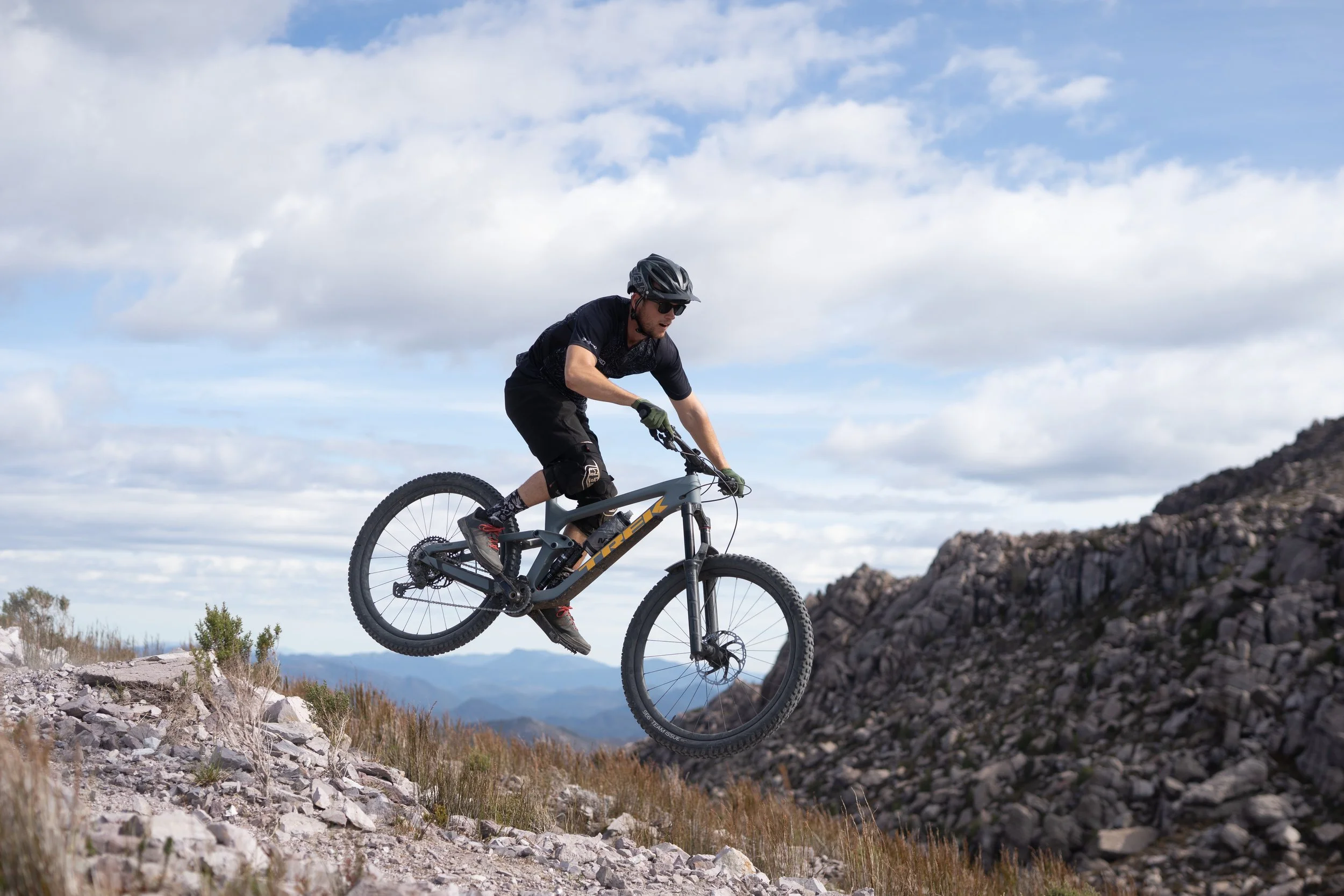 A person riding a mountain bike on rocky terrain with mountains in the background and partly cloudy sky above.