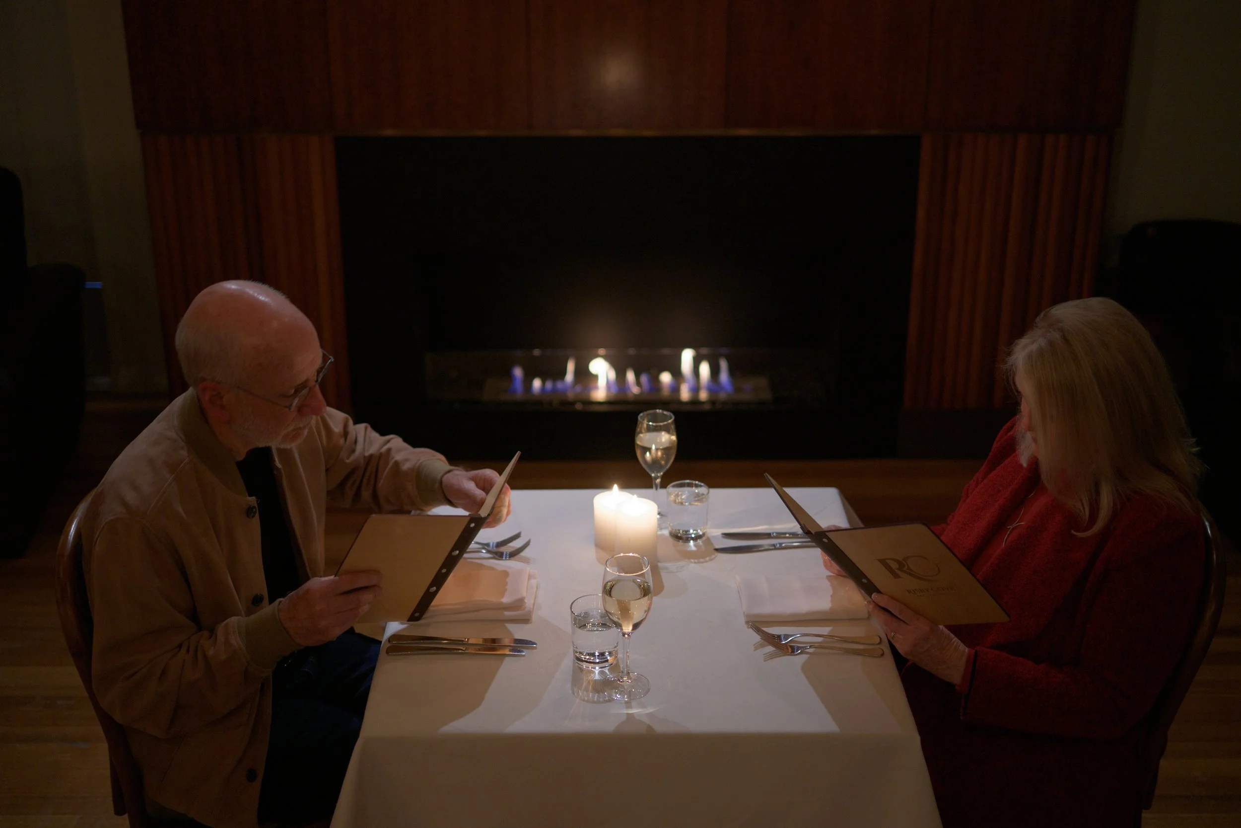 People sitting at a private dining table in the Mill at Risby Cove, reading menus, with a candle and glasses of water on the table, fireplace in the background.