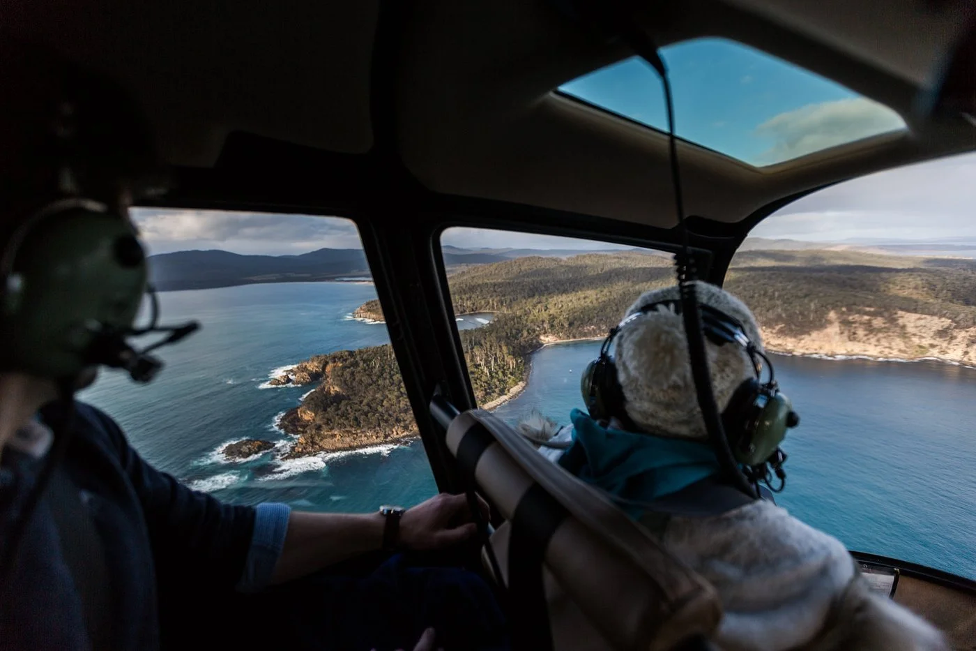 View from inside a small aircraft showing two people, one in headphones, flying over the Southern Tasmanian coastline with blue ocean and green hills.