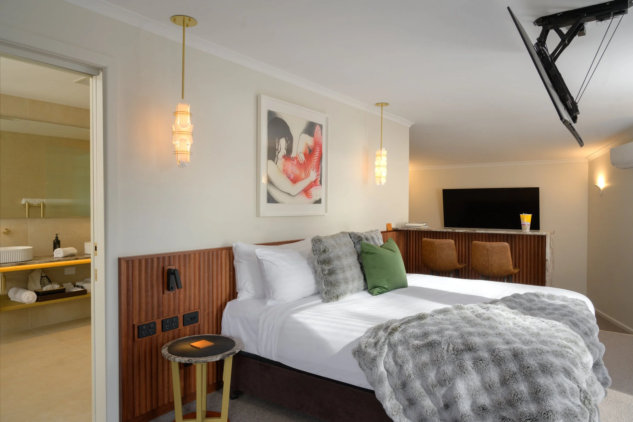 Modern hotel room with a bed, decorative pillows, artwork above the bed, a small side table, and a TV. The room has pendant lighting, neutral-colored walls, and a visible bathroom area.