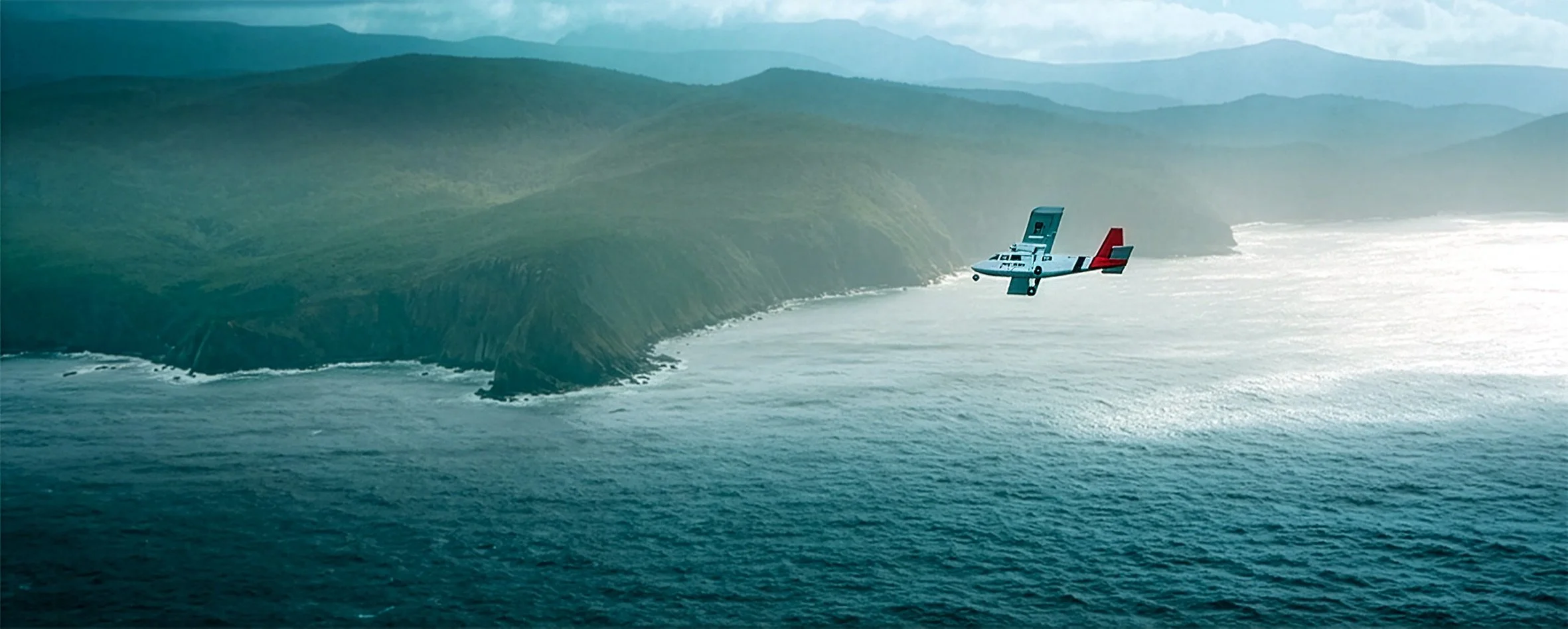A small aircraft flying over the ocean near rugged coastal cliffs with green vegetation, under a cloudy sky.