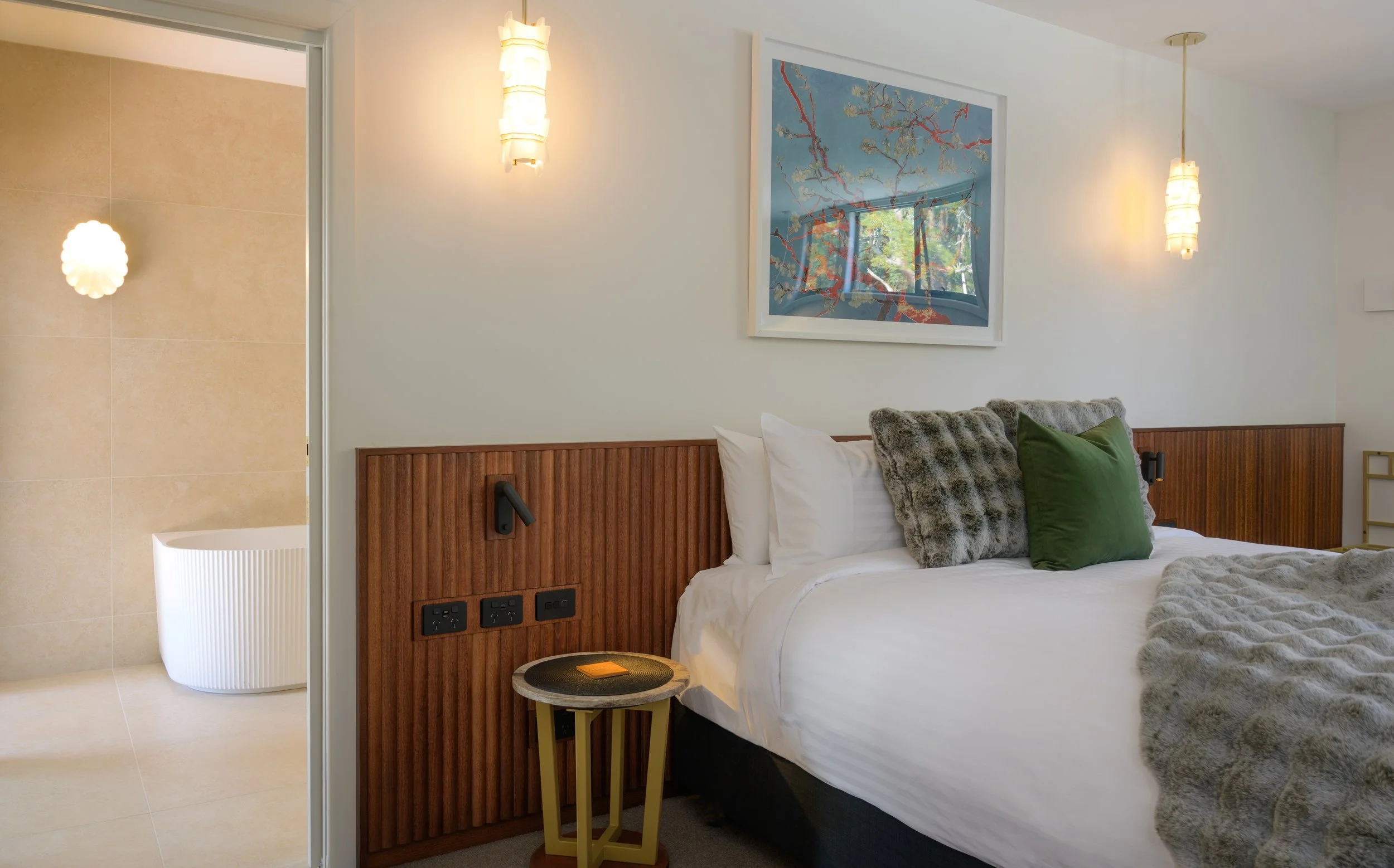 Hotel bedroom with white linens, decorative pillows, wooden headboard, framed artwork, pendant lighting, and an adjacent bathroom with beige tiles and a freestanding tub.