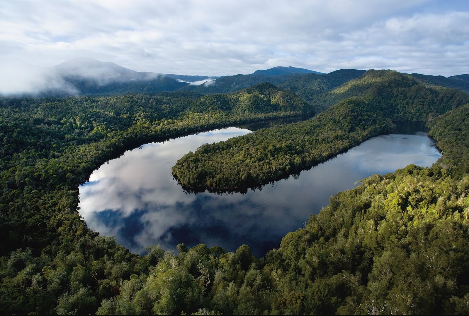 Aerial view of the Gordon River surrounded by lush green forest with mountains in the background under partly cloudy sky.