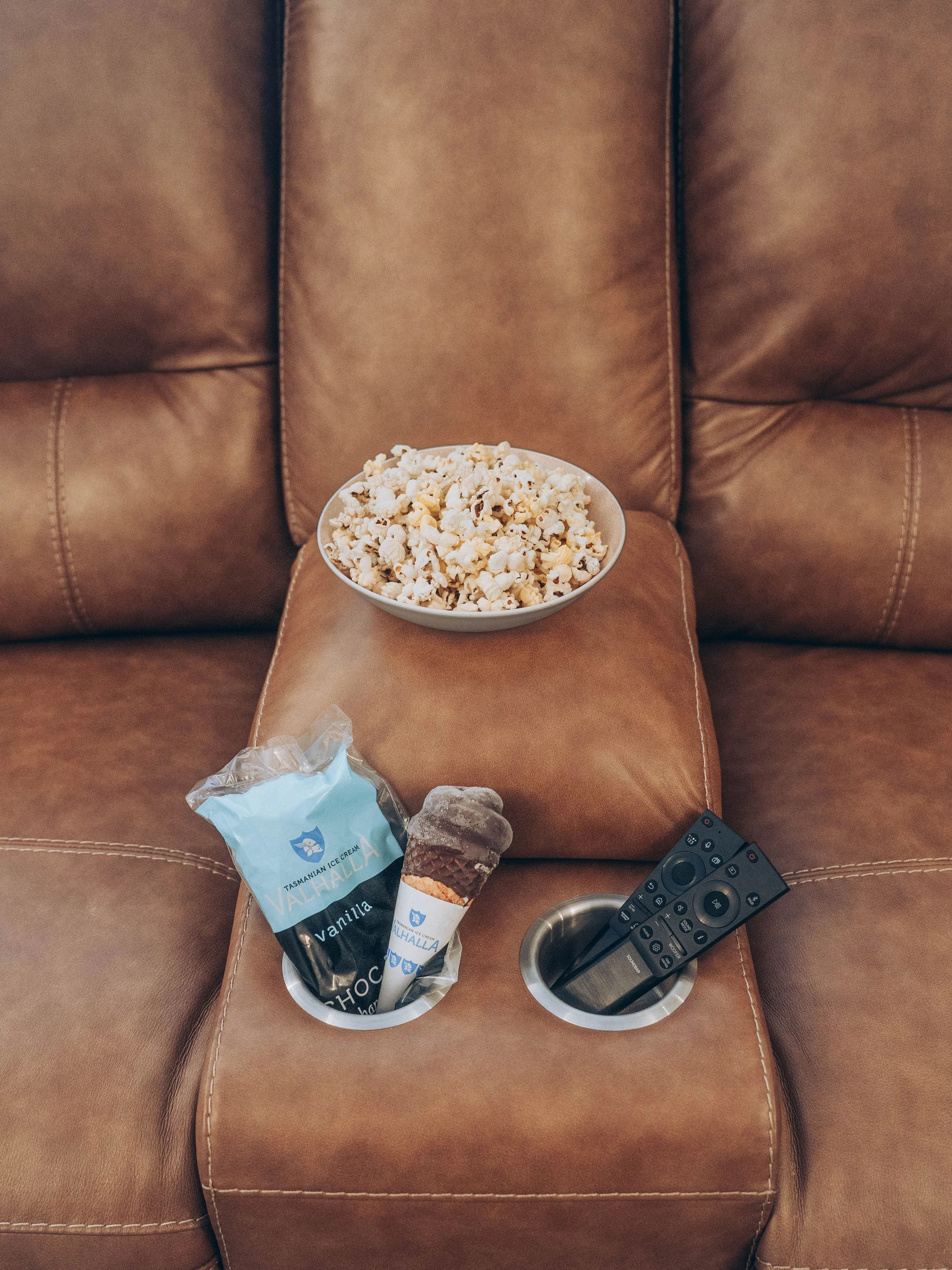 A brown leather recliner with a bowl of popcorn on the center console. The console also holds a pop-top ice cream cone and a TV remote.