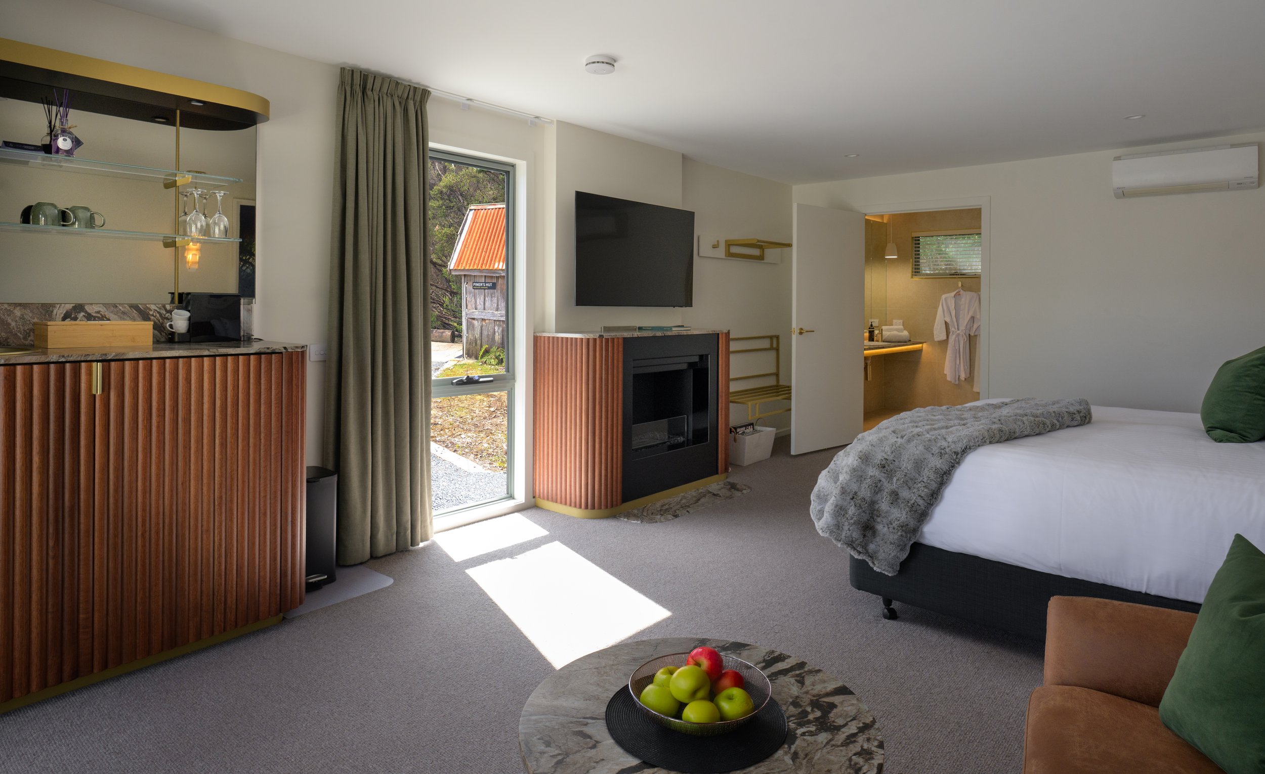 A hotel room with a large bed, a stone coffee table with fruit, a TV above a fireplace, standing mirror, and an open bathroom door with a bathrobe, with natural light coming in through the window and balcony door.
