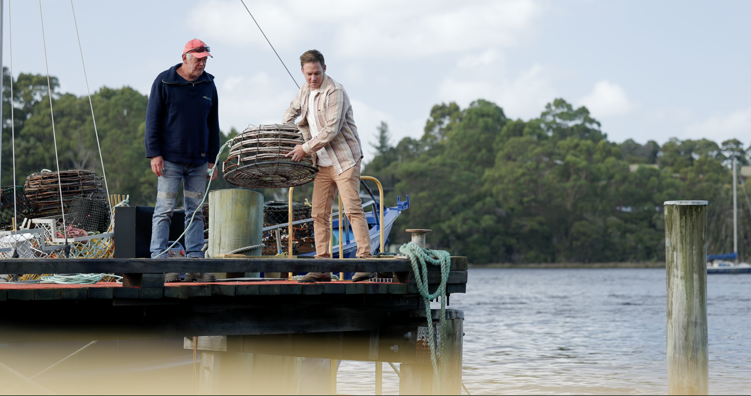 A chef standing outdoors near a body of water, holding two cooked lobsters, one in each hand, with trees and boats in the background.