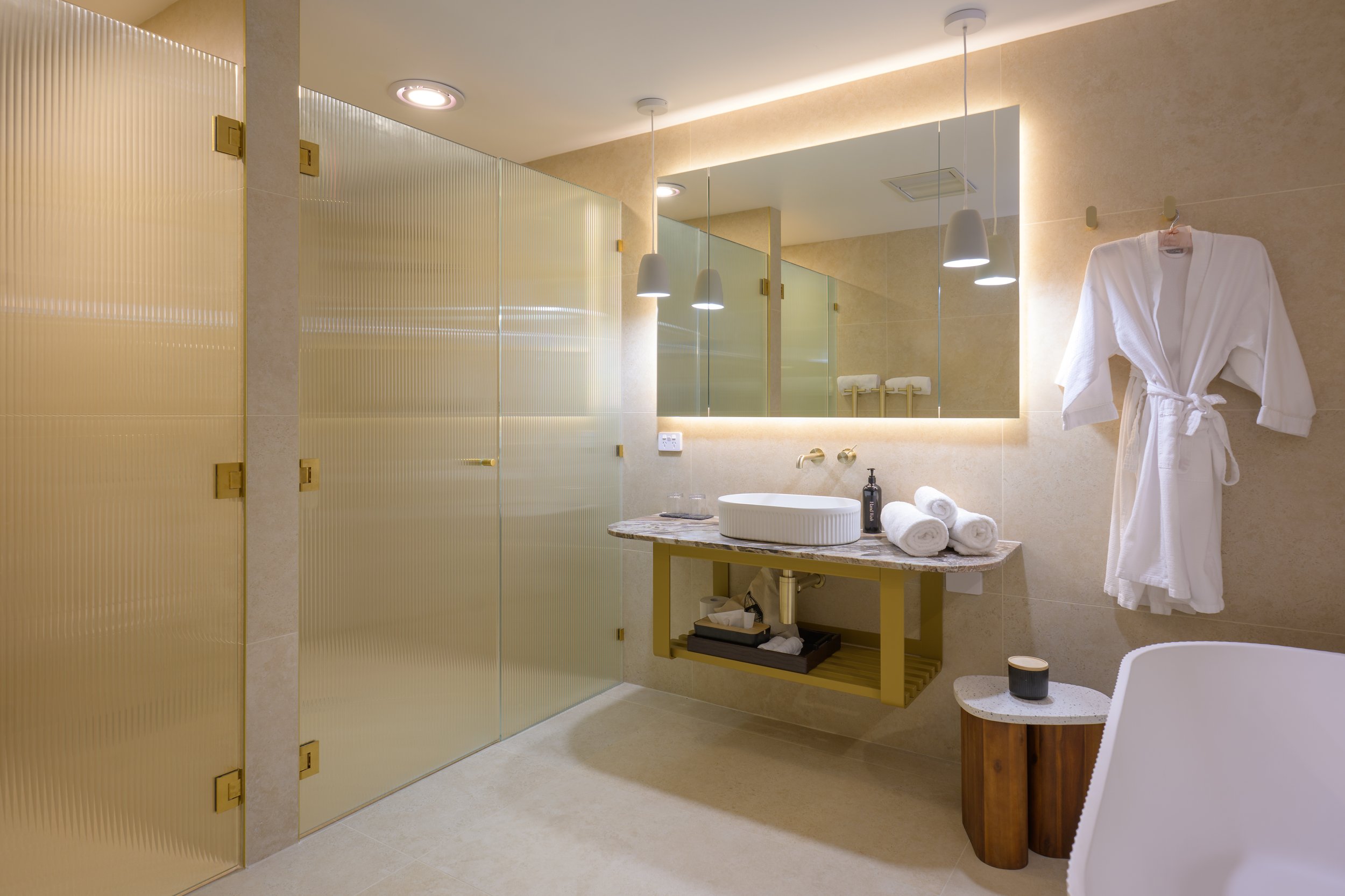 Modern bathroom with frosted glass shower enclosure, large illuminated mirror, stone countertop with sink, rolled towels, robe, small side table, and stylish lighting.