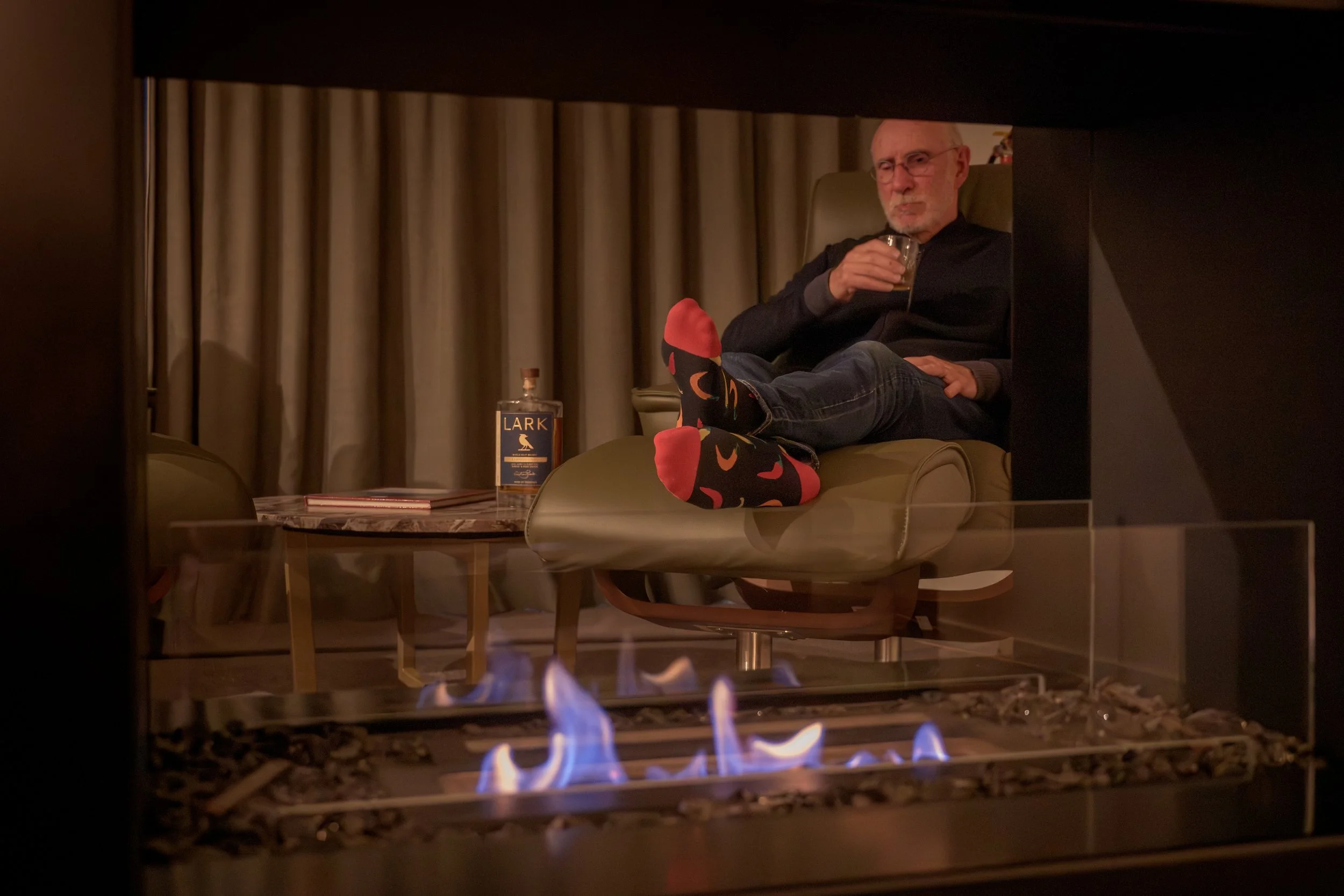 Man sitting in a chair with legs crossed, wearing colorful socks, drinking a beverage, beside a bottle of Lark whiskey, with a fireplace in the foreground, and beige curtains in the background.