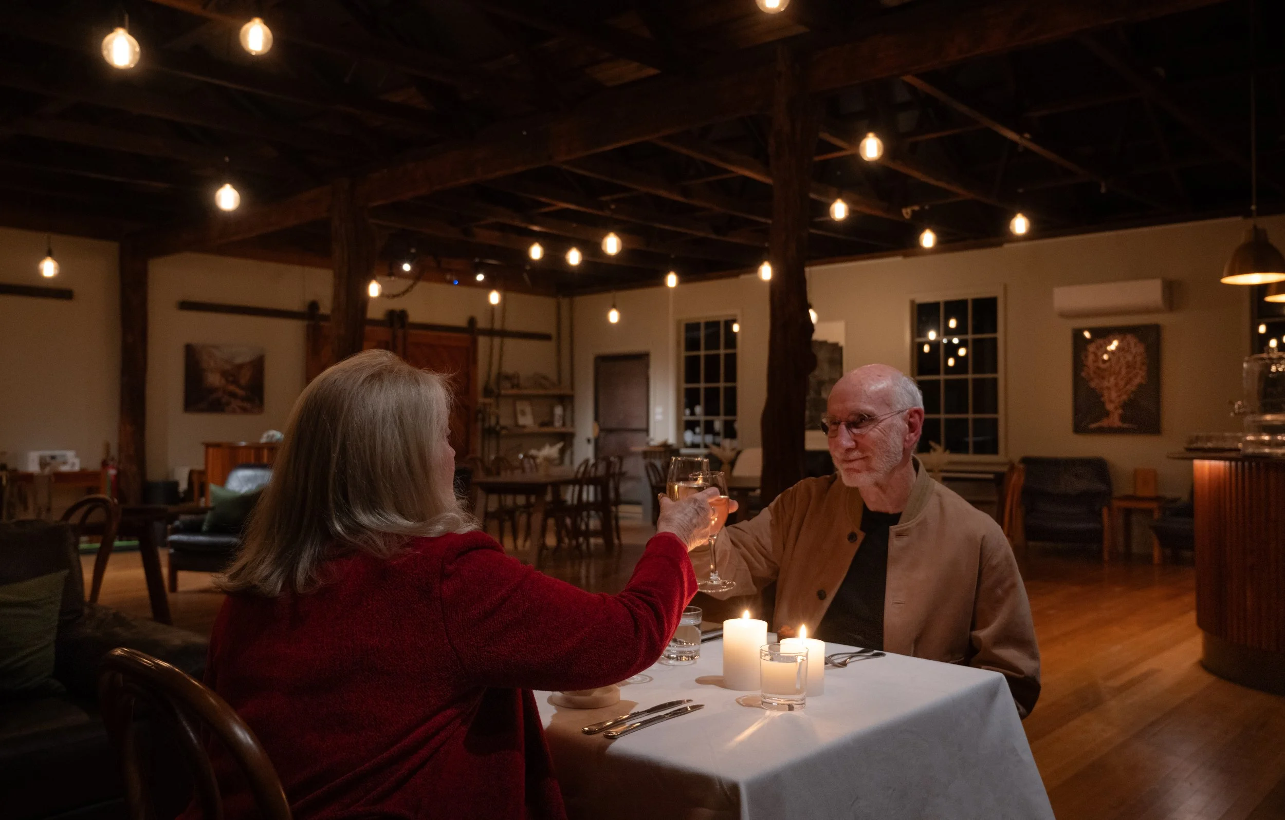 A couple have a private dinner in The Mill at Risby Cove, sitting at a table with dim lighting, candles, and warm decor. There are candles on the table and hanging light bulbs from the ceiling, creating an intimate atmosphere.