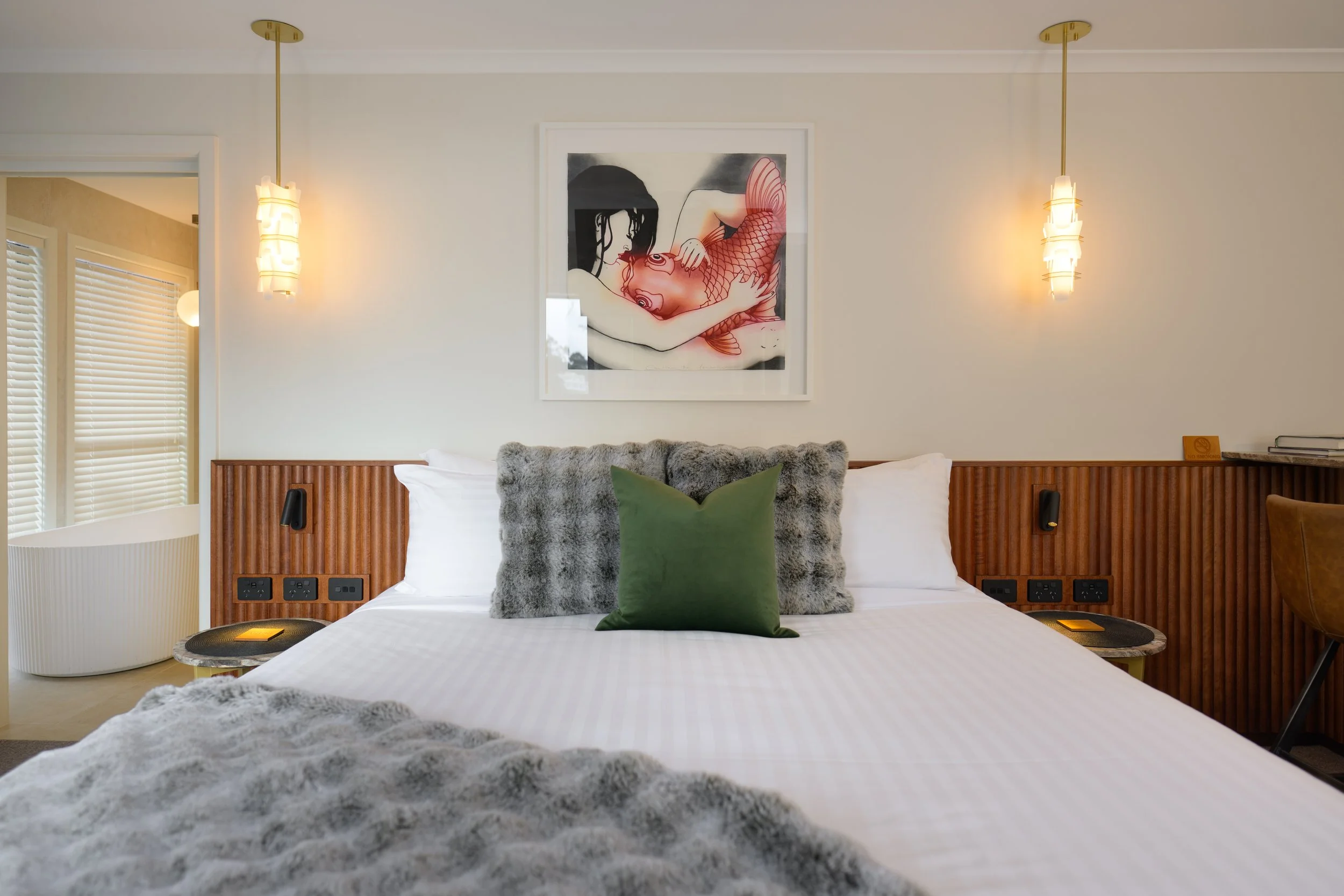 Modern hotel bedroom with a bed, gray furry pillows, and green accent pillow. Wooden headboard with built-in outlets, two hanging pendant lights, and framed wall art above the bed depicting a woman and a fish.